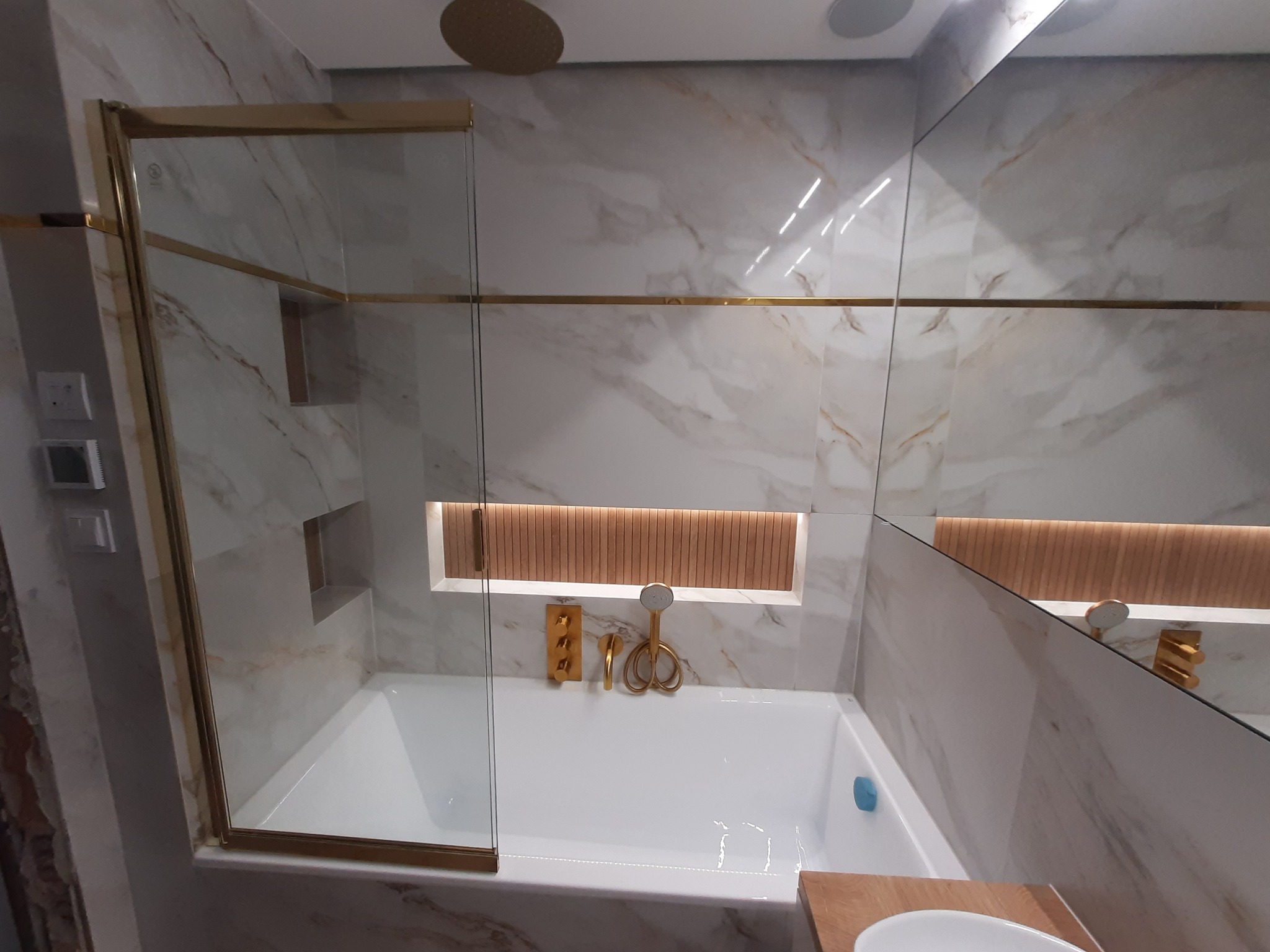Wykończona łazienka z wanną, kabiną prysznicową ze złotymi ramami, marmurowymi płytkami i złotą armaturą, oświetlona wnęka nad wanną.