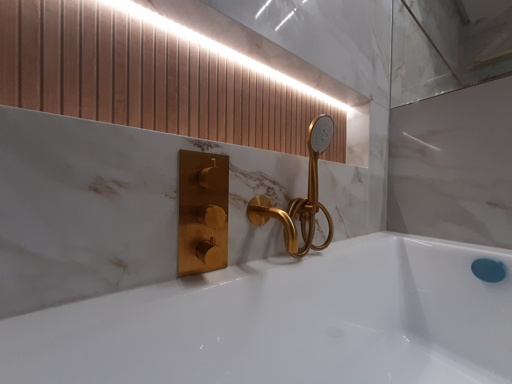 Nowoczesna łazienka z wanną, baterią i słuchawką prysznicową w kolorze szczotkowanego złota, marmurowe płytki na ścianach, oświetlenie LED w niszy z drewnianymi panelami.