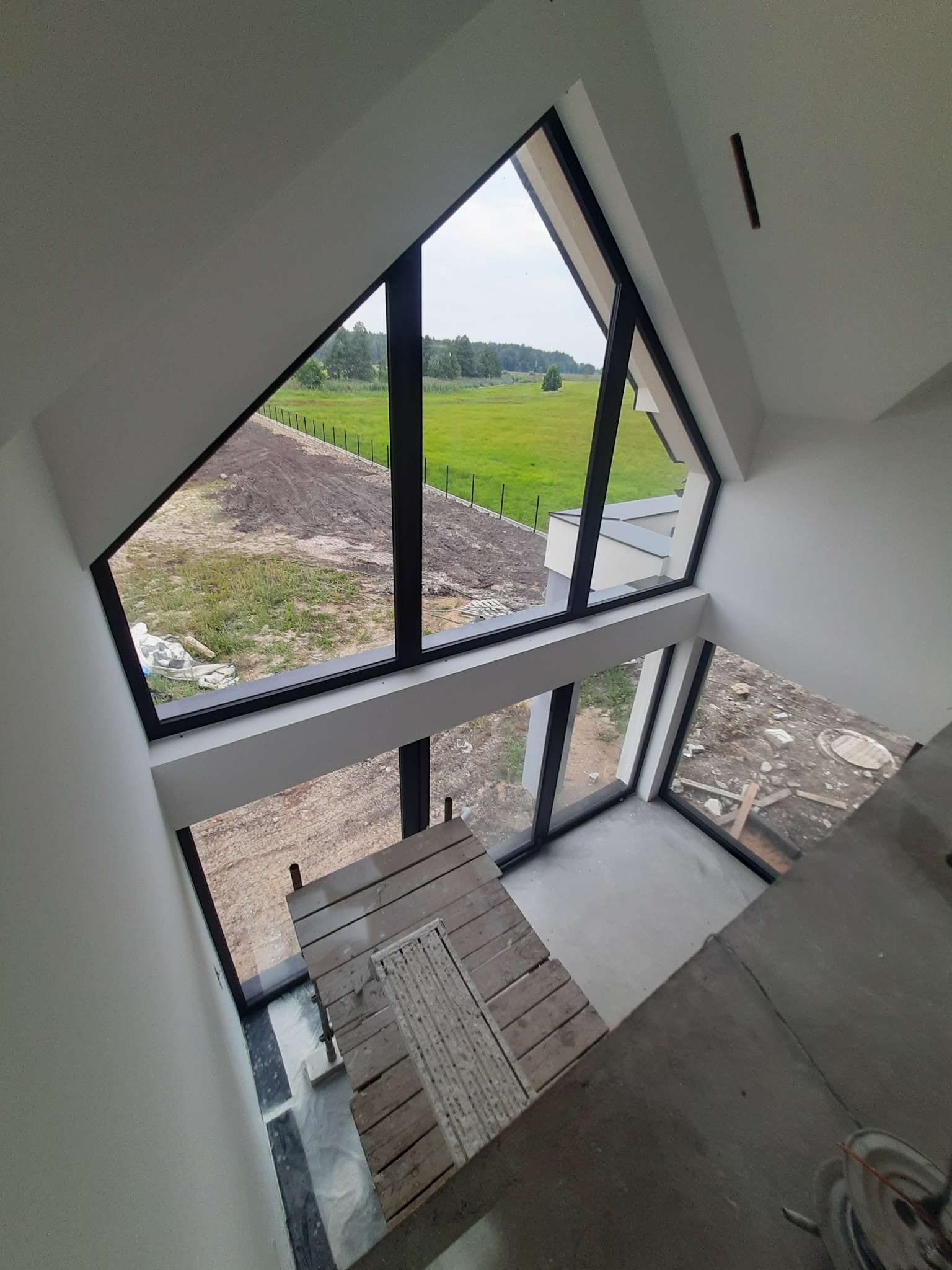 Wysokie okno w kształcie trójkąta z czarnymi ramami w domu w Augustowie, widok na zewnątrz na zielone pole i las, wewnątrz rusztowanie malarskie.
