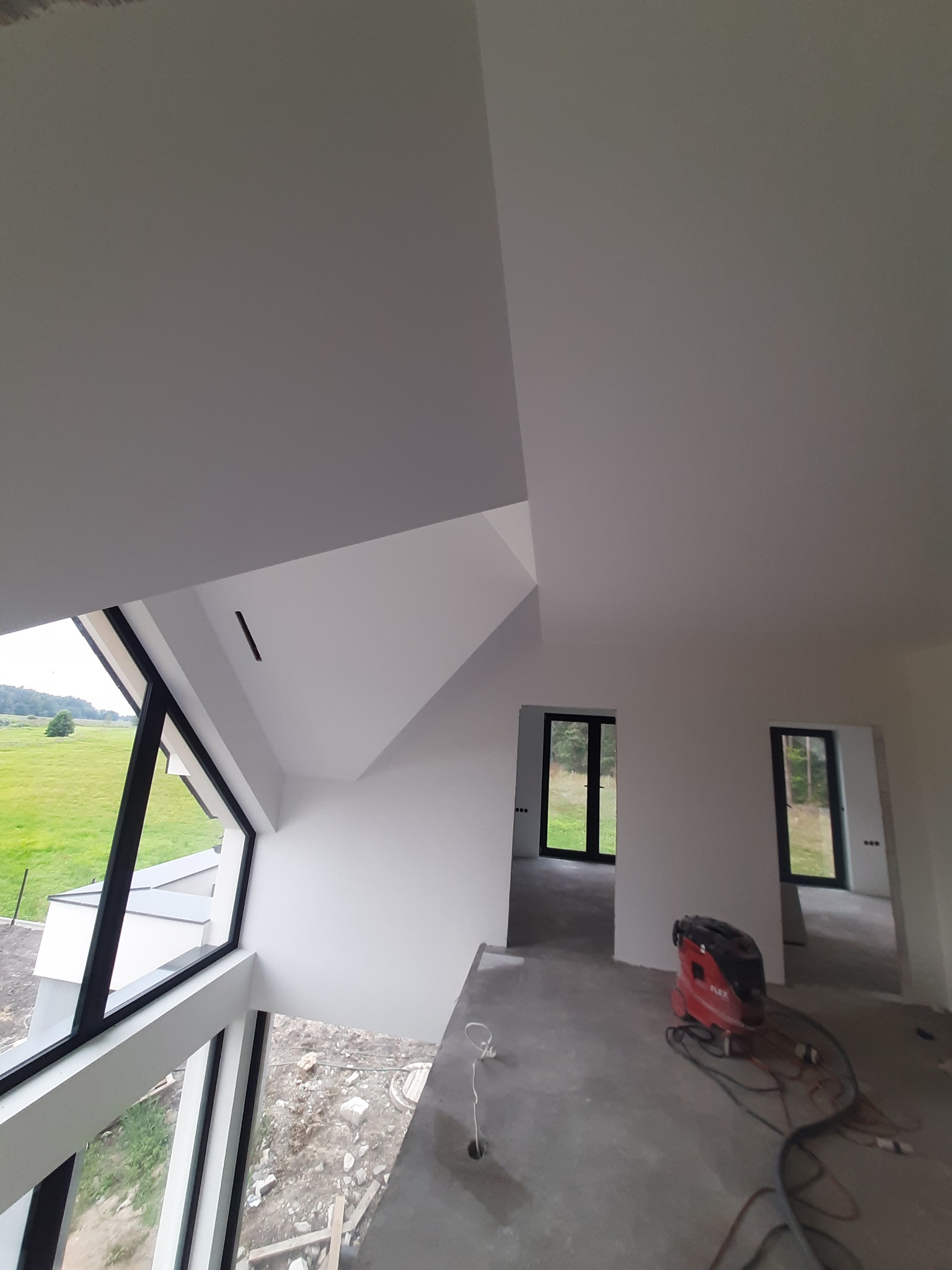Wnętrze domu w trakcie wykańczania, widoczne duże okna z widokiem na zieleń, białe ściany i sufit, szara podłoga oraz czerwony odkurzacz przemysłowy.