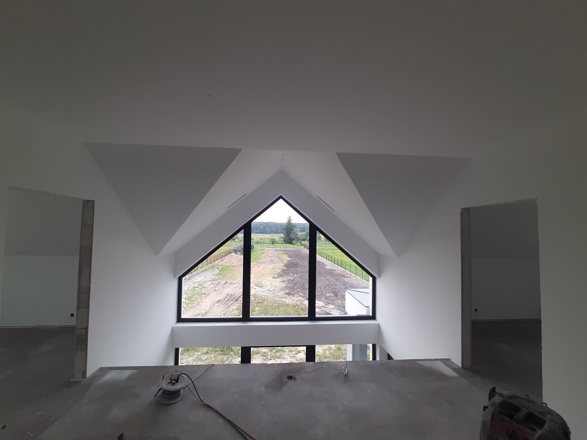 Wnętrze poddasza z dużym trójkątnym oknem z widokiem na zewnątrz, surowy beton na podłodze, świeżo pomalowane ściany na biało, widoczne kable i narzędzia sugerujące prace wykończeniowe.