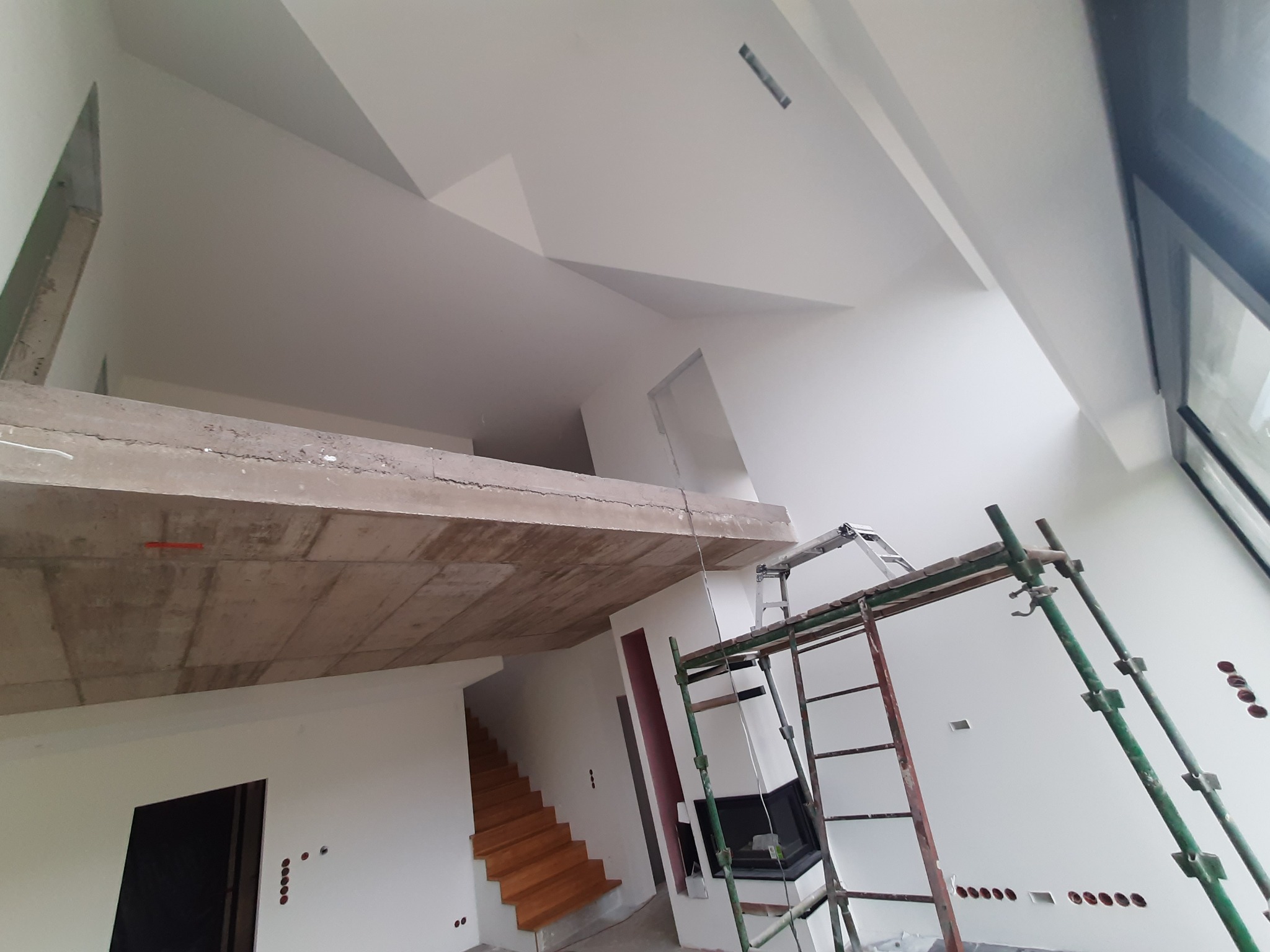 Wnętrze domu w trakcie prac wykończeniowych: białe ściany, surowy betonowy strop, drewniane schody, rusztowanie przy ścianie z przygotowanymi otworami pod gniazdka.