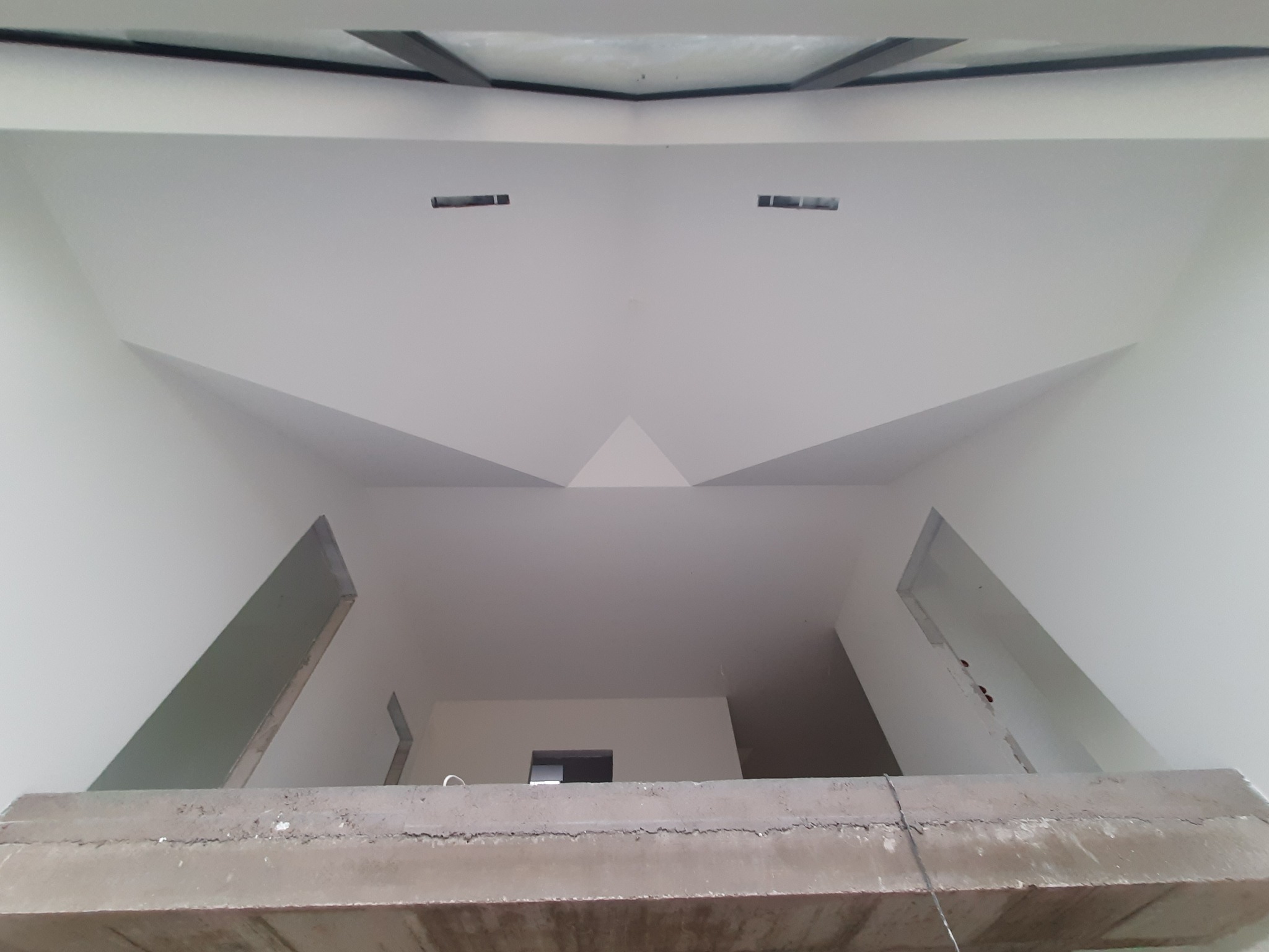 Surowe wnętrze z widocznymi ścianami po szpachlowaniu i malowaniu na biało, z fragmentem betonowej konstrukcji na pierwszym planie, widok z dołu na sufit z otworami na oświetlenie.