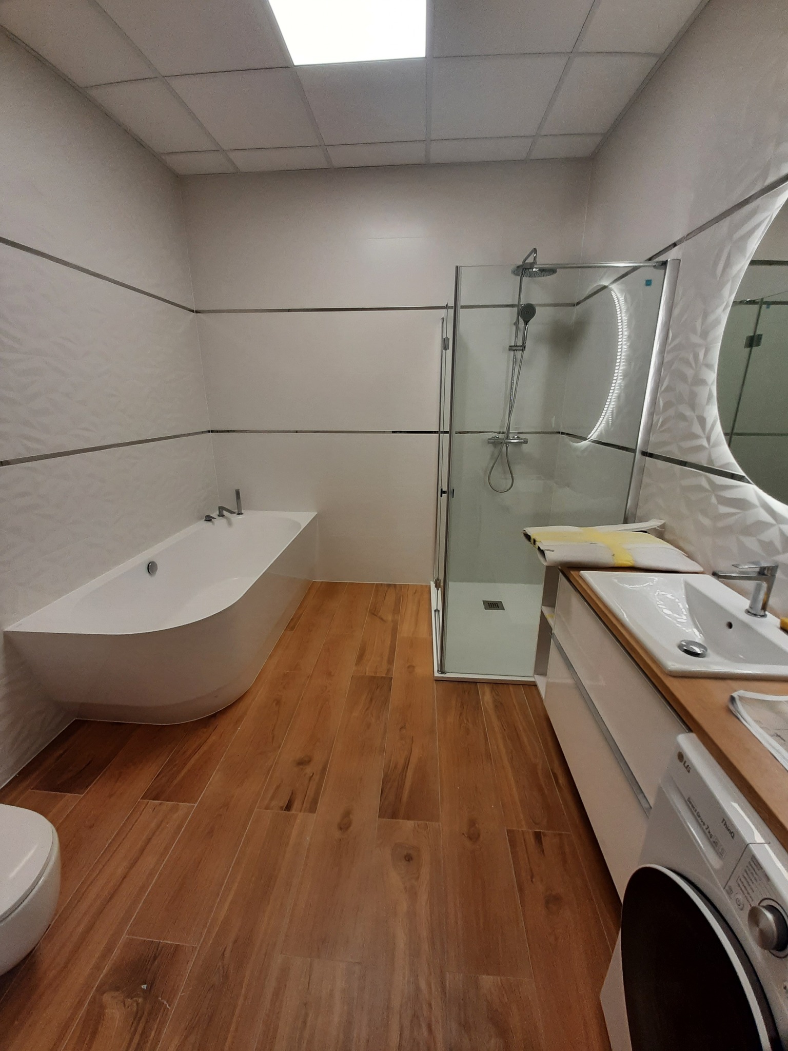 Nowoczesna łazienka z drewnopodobną podłogą, białą wanną asymetryczną, kabiną prysznicową ze szklanymi drzwiami, umywalką z drewnianym blatem i pralką LG.