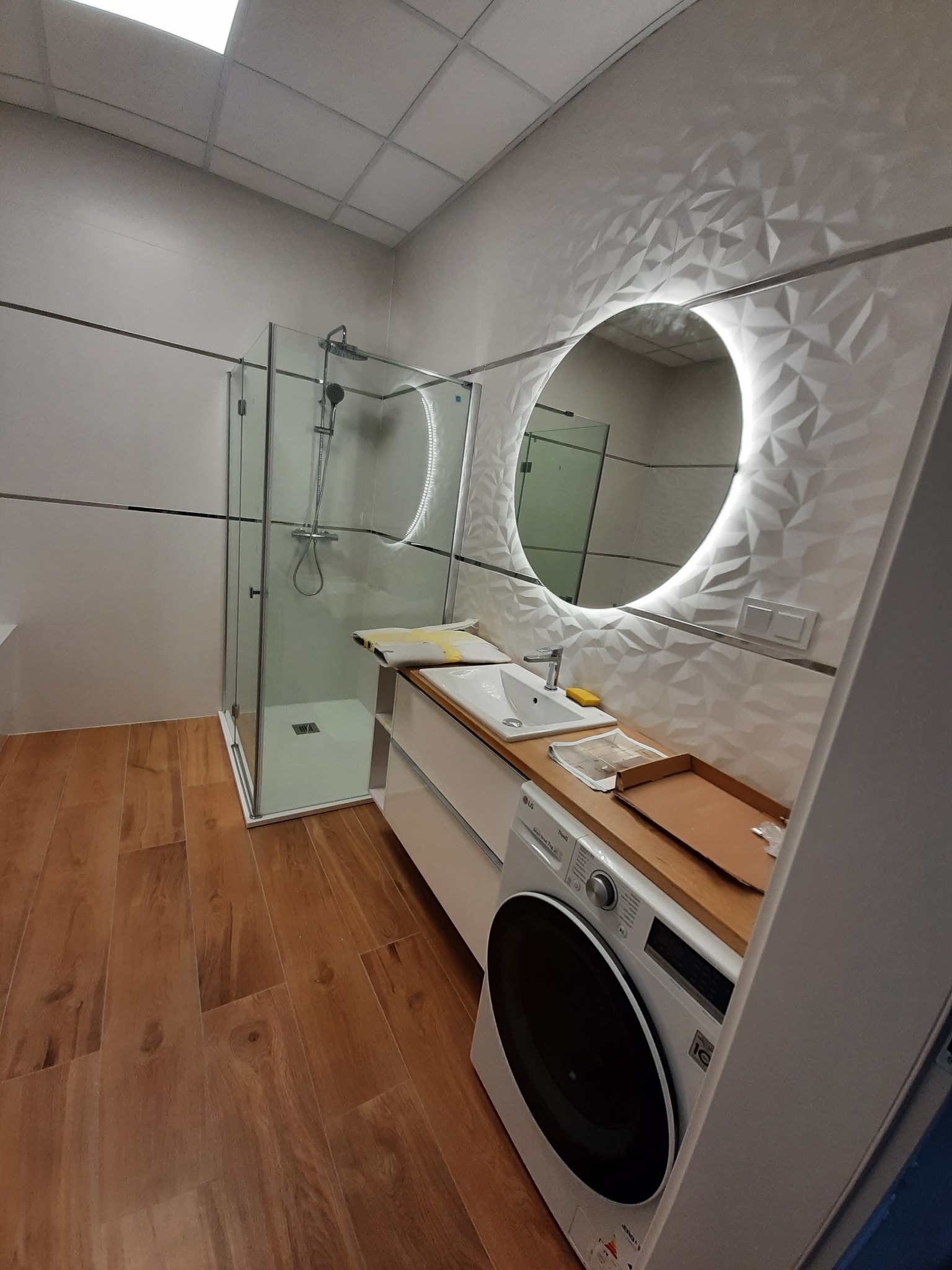 Nowoczesna łazienka z kabiną prysznicową, umywalką w zabudowie z pralką i okrągłym lustrem z podświetleniem LED na ścianie z trójwymiarową teksturą.