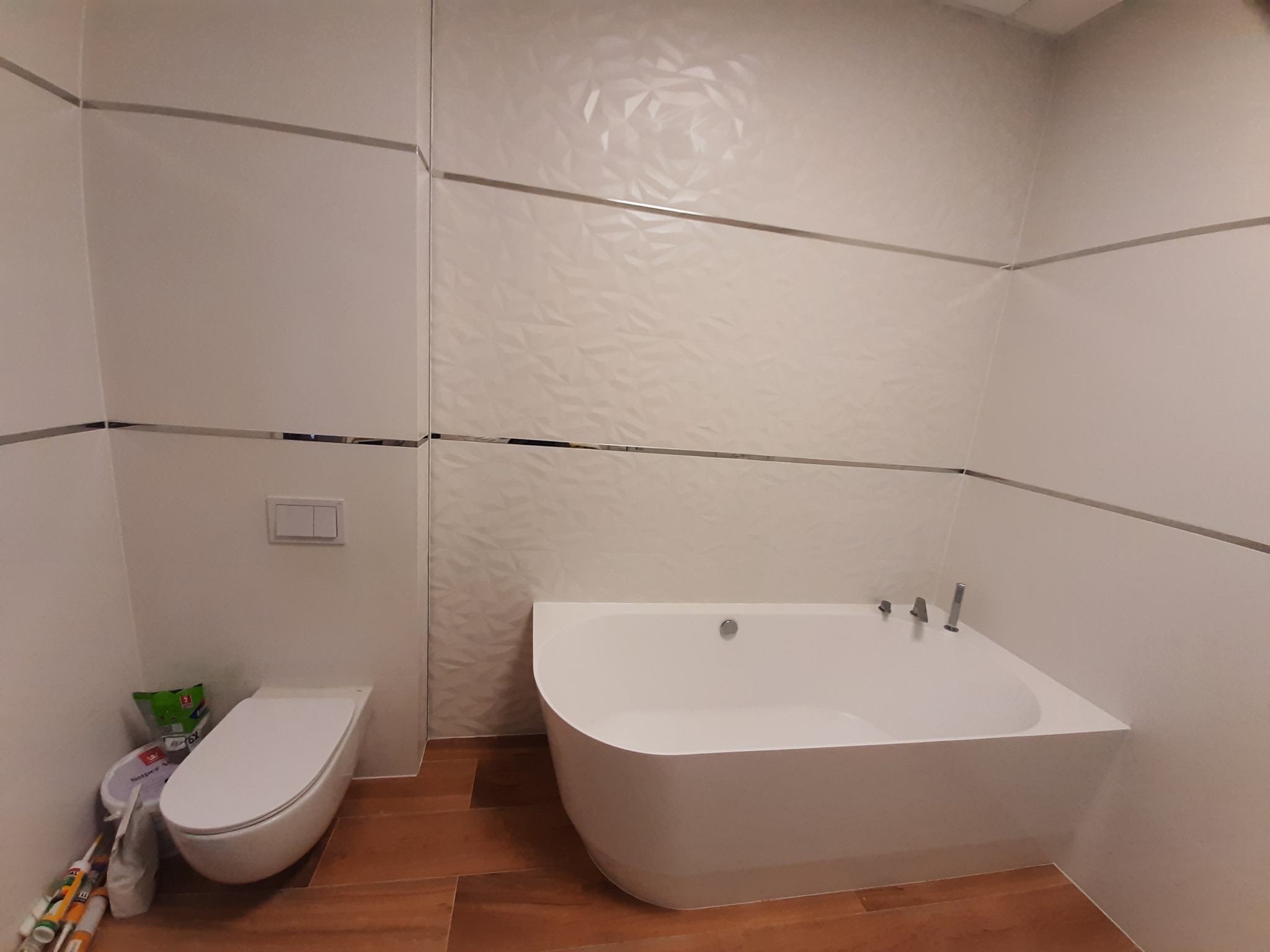 Nowoczesna łazienka z białą wanną narożną, podwieszaną toaletą i płytkami ściennymi z subtelnym wzorem 3D, wykończonymi metalowymi listwami; widoczne materiały budowlane obok toalety.