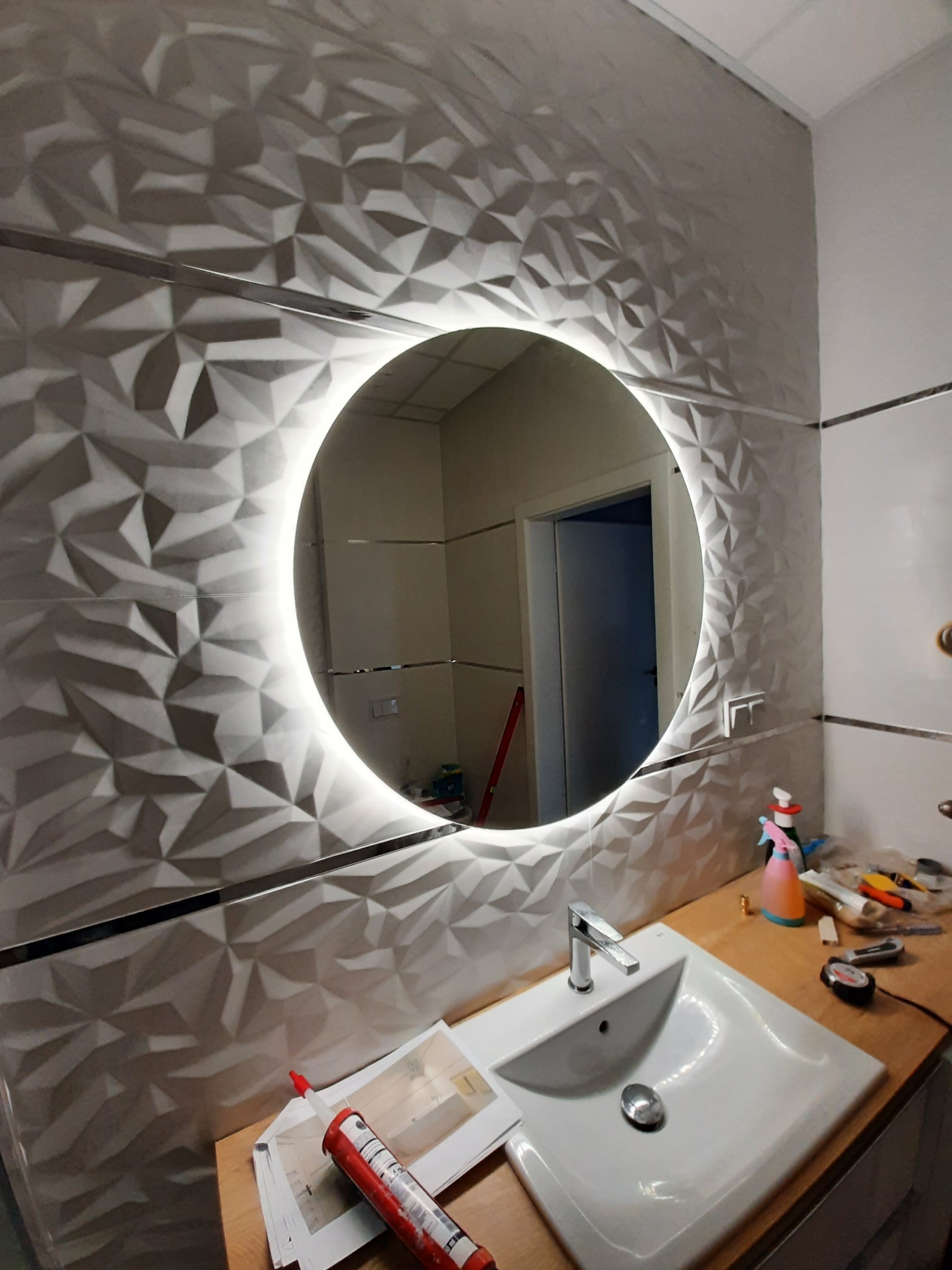 Łazienka po renowacji: ściana wyłożona trójwymiarowymi płytkami ceramicznymi w odcieniach szarości, okrągłe lustro podświetlane LED, biała umywalka z chromowaną baterią, widoczne narzędzia...