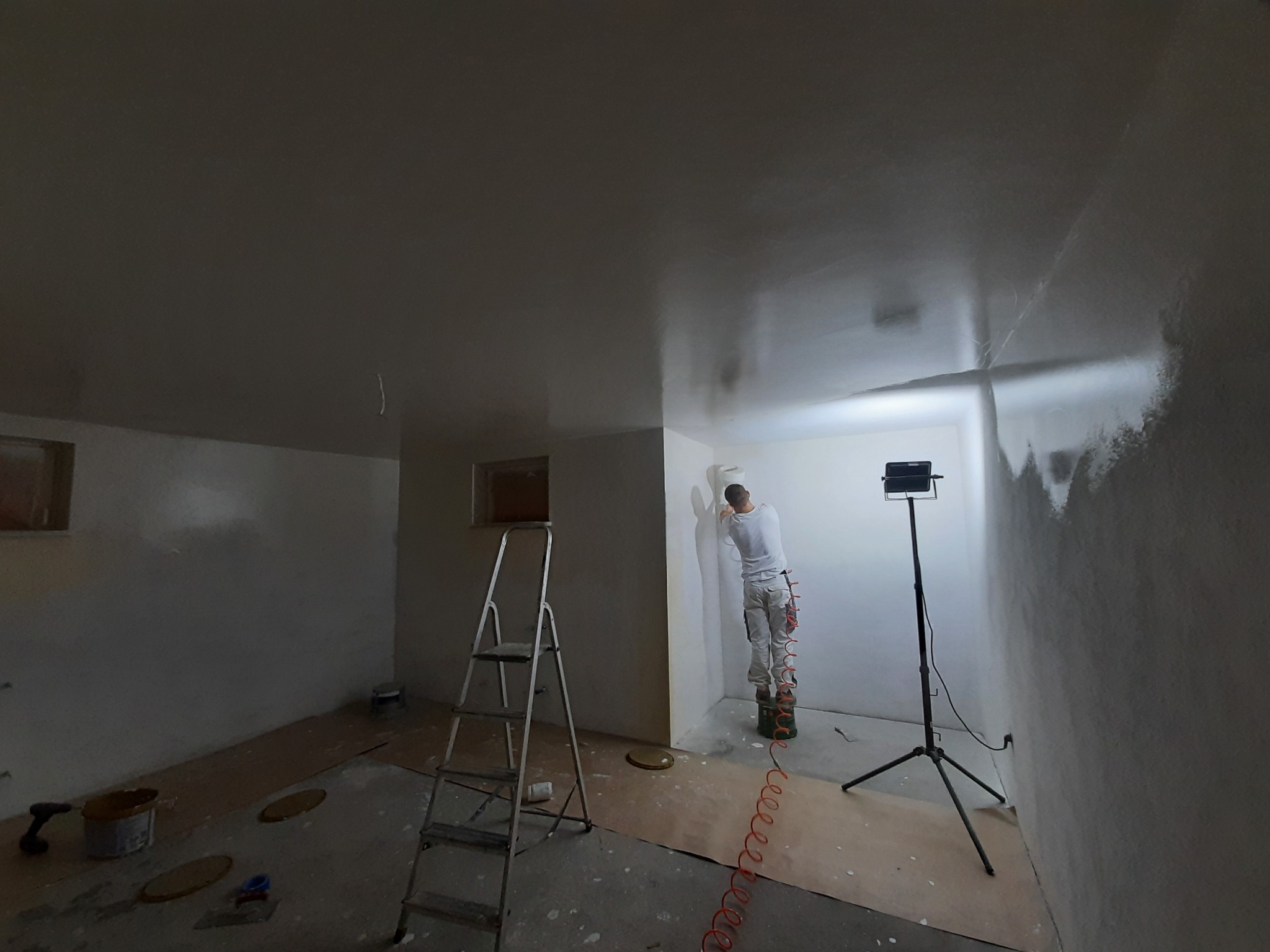 Mężczyzna szpachluje ścianę w pokoju, stojąc na podwyższeniu, z widoczną drabiną, lampą oświetleniową i narzędziami malarskimi.