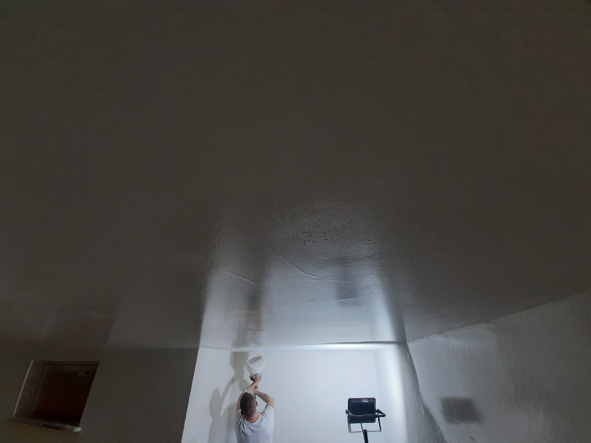 Praca na wysokości: mężczyzna szpachluje sufit w pomieszczeniu oświetlonym halogenem, widoczna faktura świeżo nałożonej masy.