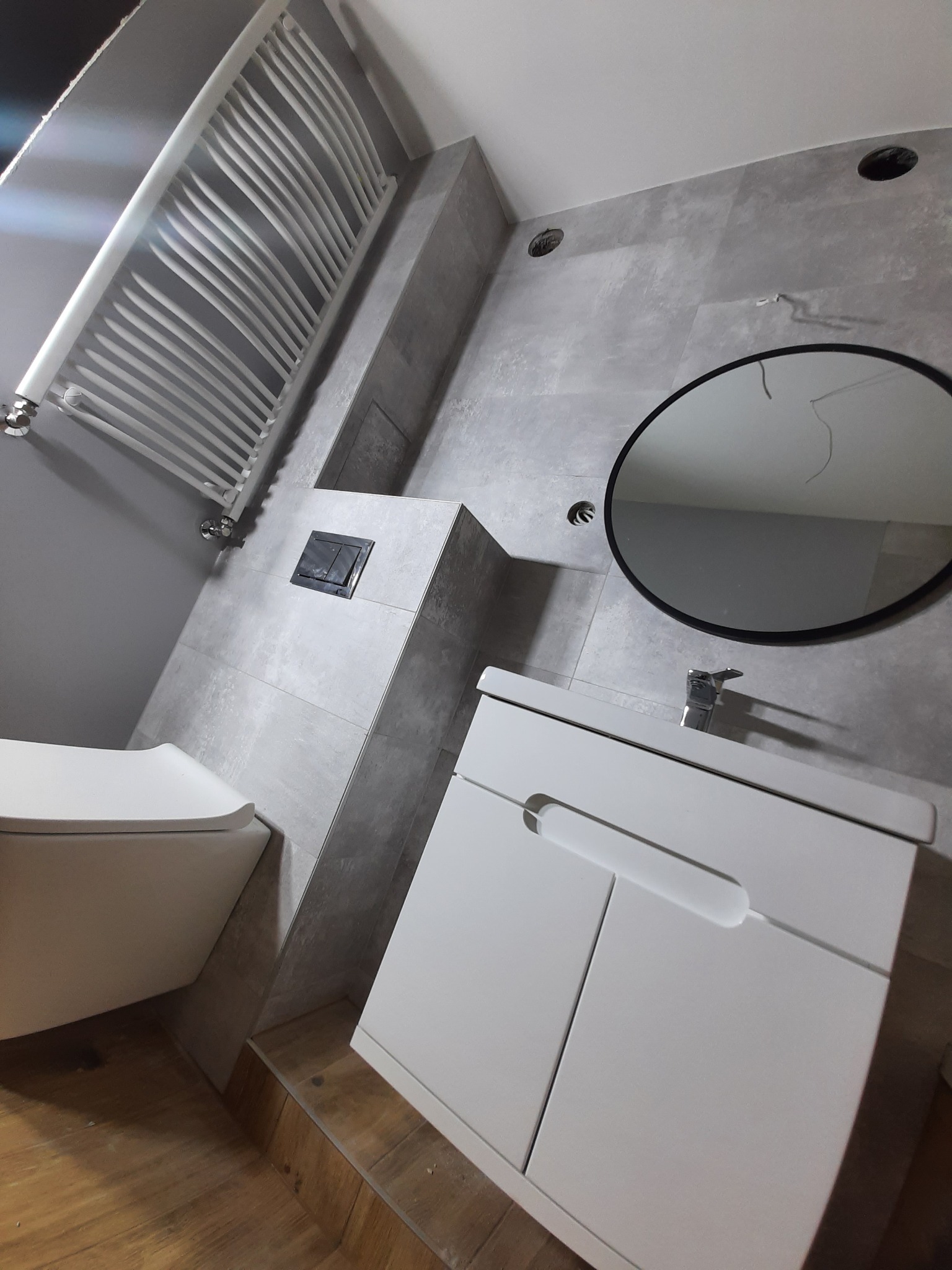 Wykończona łazienka z szarymi płytkami, wiszącą toaletą, białym grzejnikiem drabinkowym i szafką pod umywalką z okrągłym lustrem.
