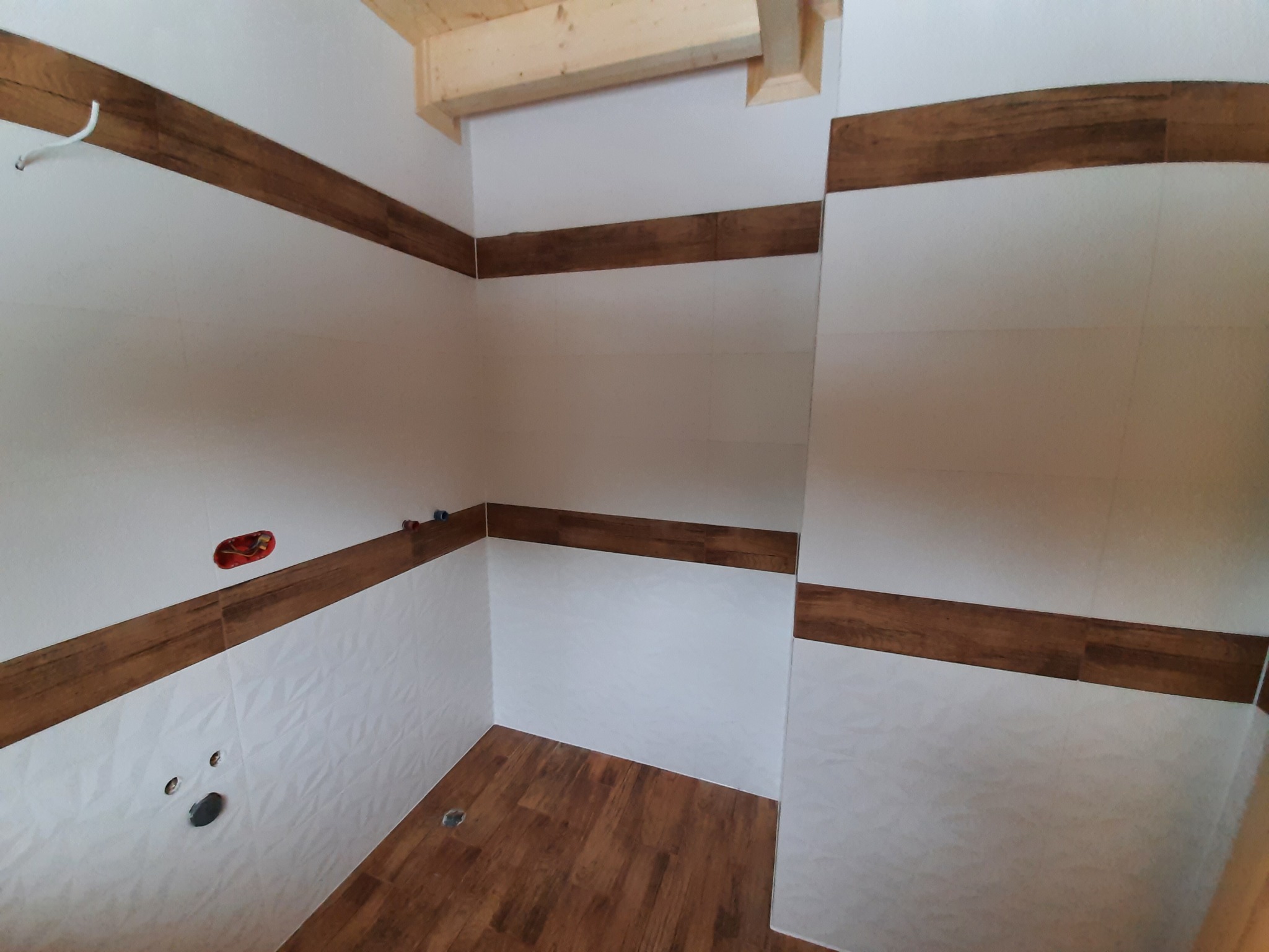 Wnętrze łazienki z częściowo ułożonymi białymi płytkami z delikatnym wzorem i pasem brązowych płytek imitujących drewno, widoczne otwory na instalacje i fragment drewnianej konstrukcji sufitu.