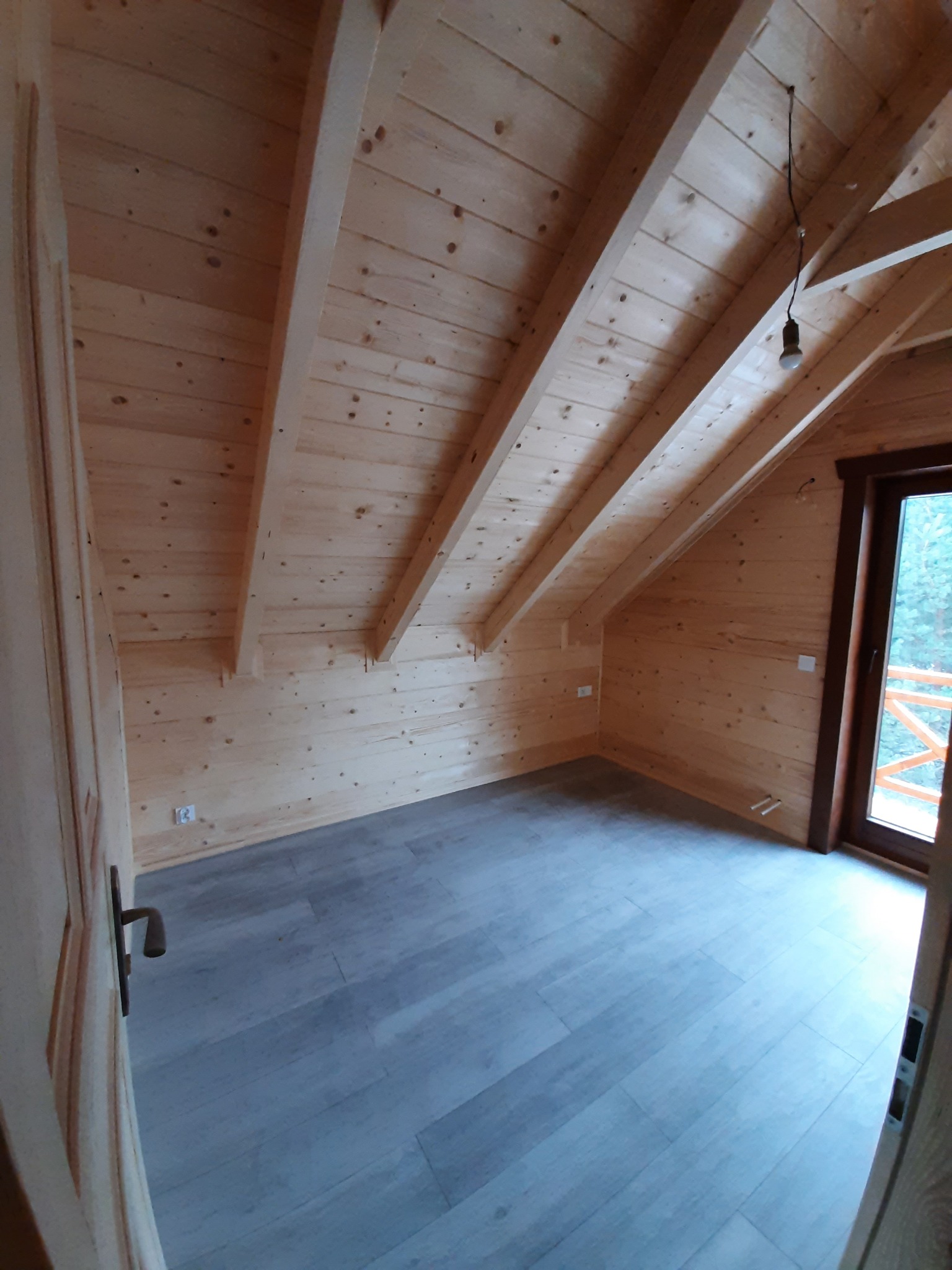 Poddasze wykończone boazerią, z drewnianymi belkami stropowymi i szarą podłogą z paneli, widok z otwartych drzwi na balkon.