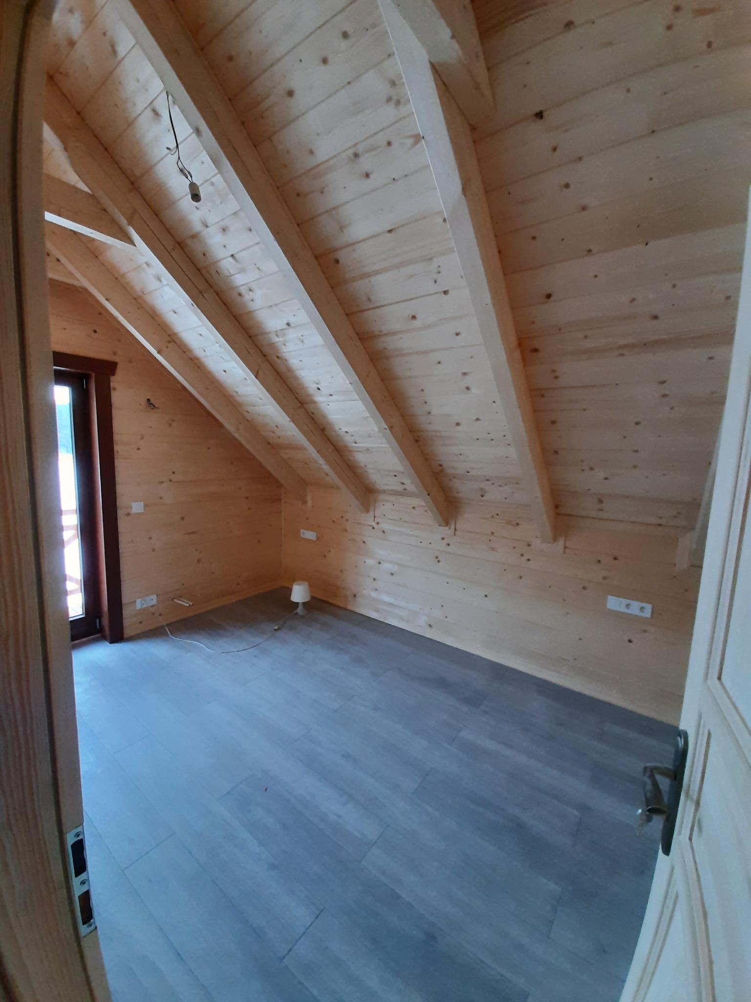 Poddasze wykończone drewnem z szarymi panelami podłogowymi, widok zza uchylonych drzwi, widoczne skosy, gniazdka elektryczne i minimalistyczna lampka.