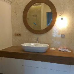 Łazienka z okrągłym lustrem w drewnianej ramie, białą umywalką nablatową i blatem z drewna na białych szafkach; widoczne gniazdka elektryczne w ścianie z beżowych płytek z kwiatowym wzorem.