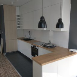 Viktor Hanzii - Nowoczesna kuchnia z białymi frontami i drewnianym blatem, czarnym zlewem i wiszącymi lampami, widok na fragment podłogi z płytek i paneli.