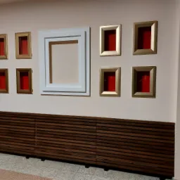 Drewniana zabudowa grzejnika z poziomych listew, nad nią rząd pustych ramek w odcieniach złota i bieli na beżowej ścianie.