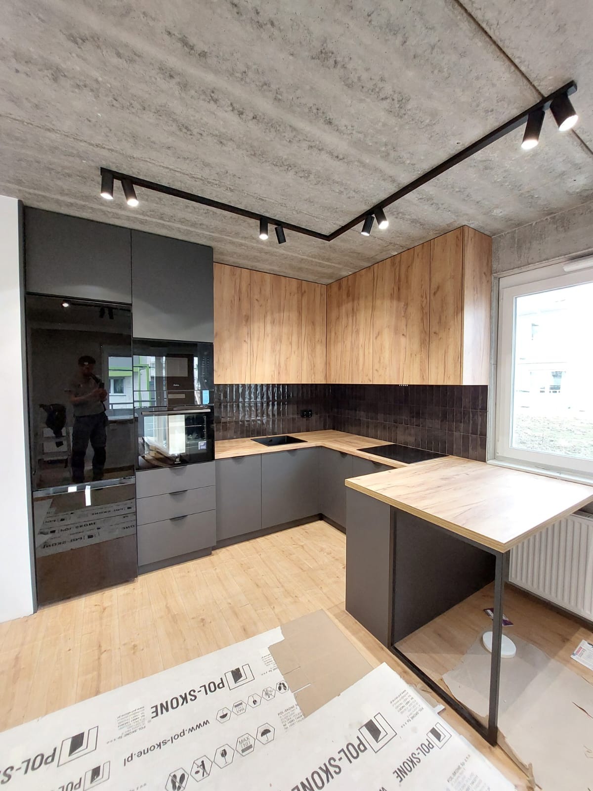 Nowoczesna kuchnia z szarymi szafkami i drewnopodobnymi frontami górnymi, czarnymi płytkami nad blatem i betonowym sufitem z oświetleniem punktowym, z widocznymi kartonami ochronnymi na podłodze.