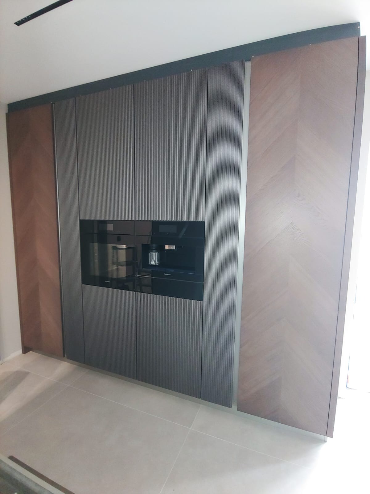 Nowoczesna zabudowa kuchenna z ciemnoszarymi ryflowanymi frontami i wbudowanym ekspresem do kawy oraz piekarnikiem Miele, boki wykończone drewnem w jodełkę, jasna podłoga.