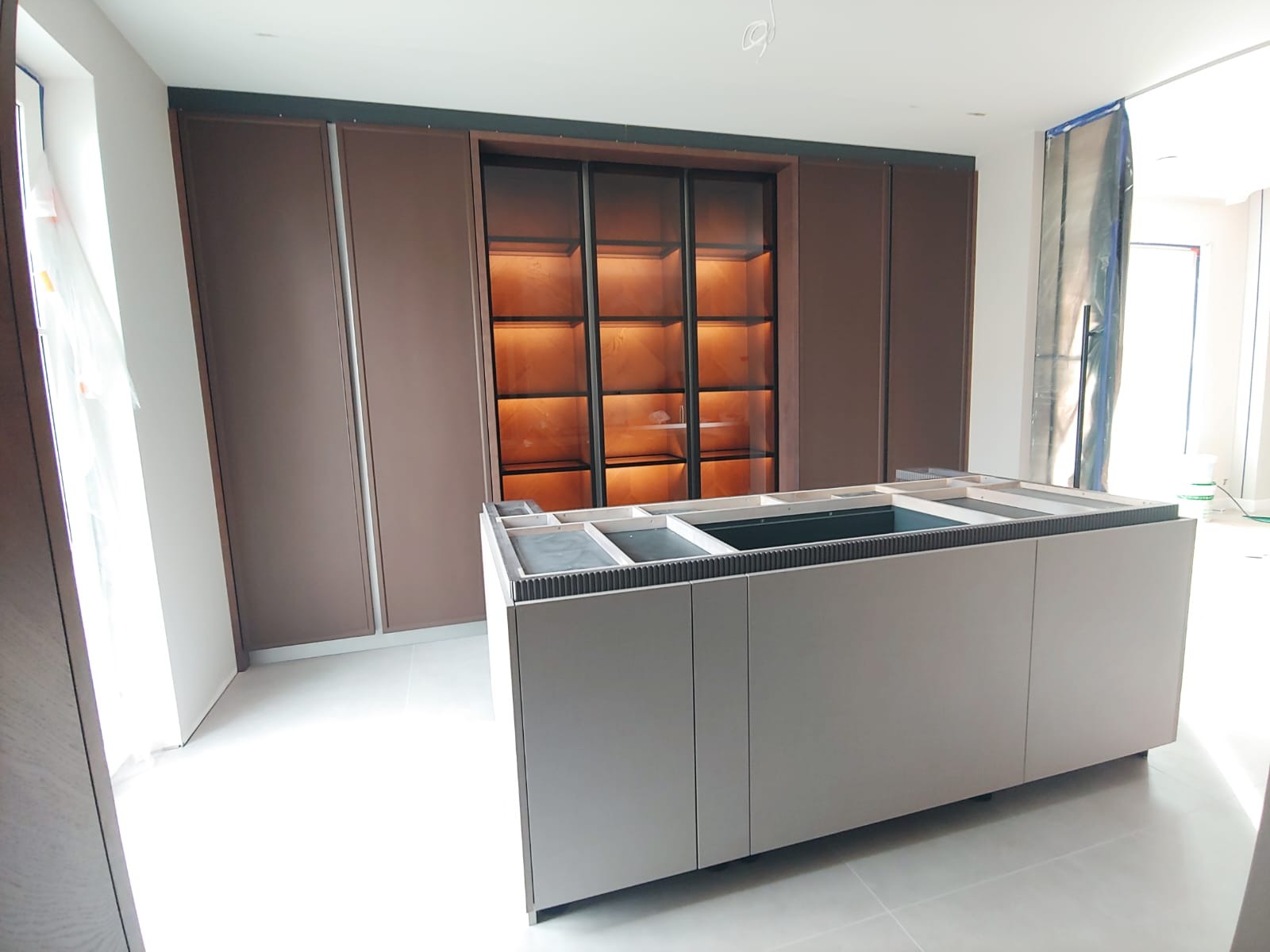 Wnętrze w trakcie montażu kuchni: widoczna wyspa kuchenna bez blatu i zabudowa ścienna z podświetlanymi półkami w tle.