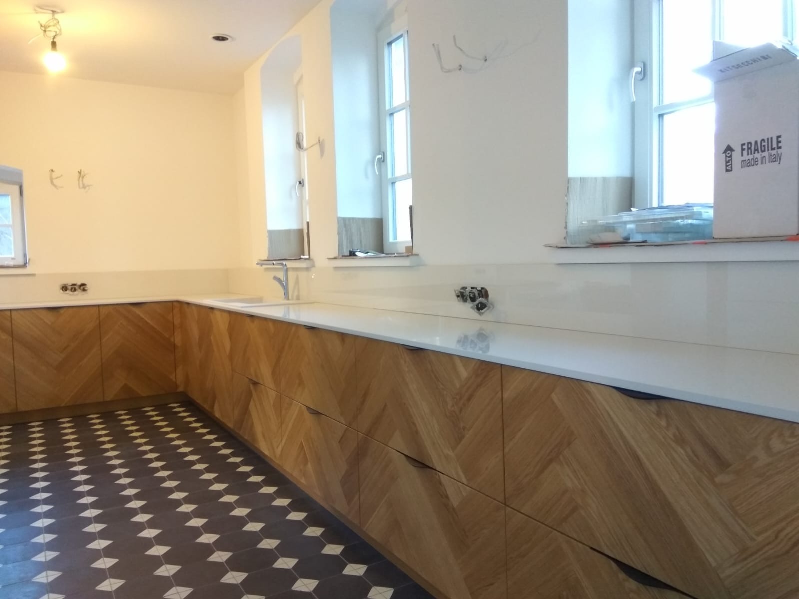 Jasna kuchnia z drewnianymi szafkami ułożonymi w jodełkę, białym blatem i podłogą w czarno-białą szachownicę. Widoczne gniazdka elektryczne nad blatem oraz kartonowe pudło przy oknie.