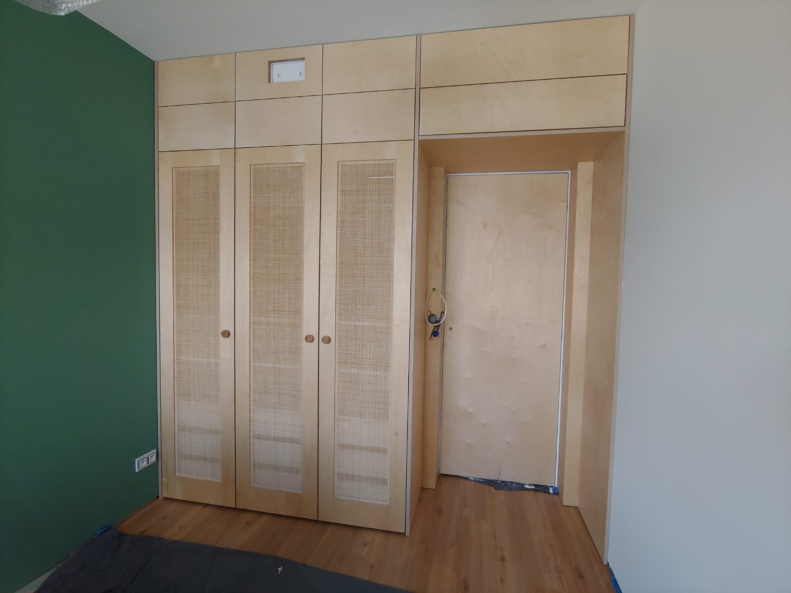 Wbudowana szafa z trzema drzwiami z rattanową wstawką, zintegrowana z drzwiami wewnętrznymi, widoczny fragment zielonej ściany i fragment podłogi laminowanej.
