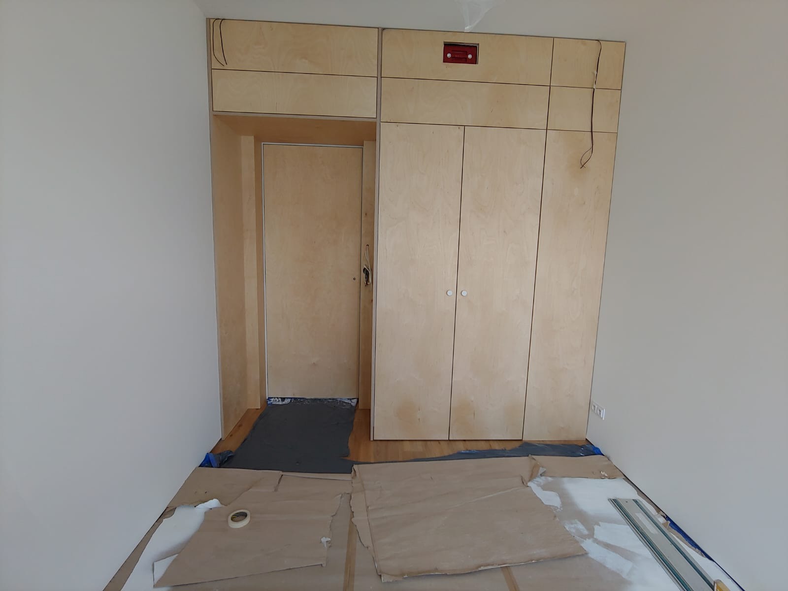 Wnętrze pokoju w trakcie montażu zabudowy z jasnej sklejki, widoczna szafa z drzwiami na zawiasach, górne szafki, otwór drzwiowy obudowany sklejką, podłoga zabezpieczona kartonem i folią.