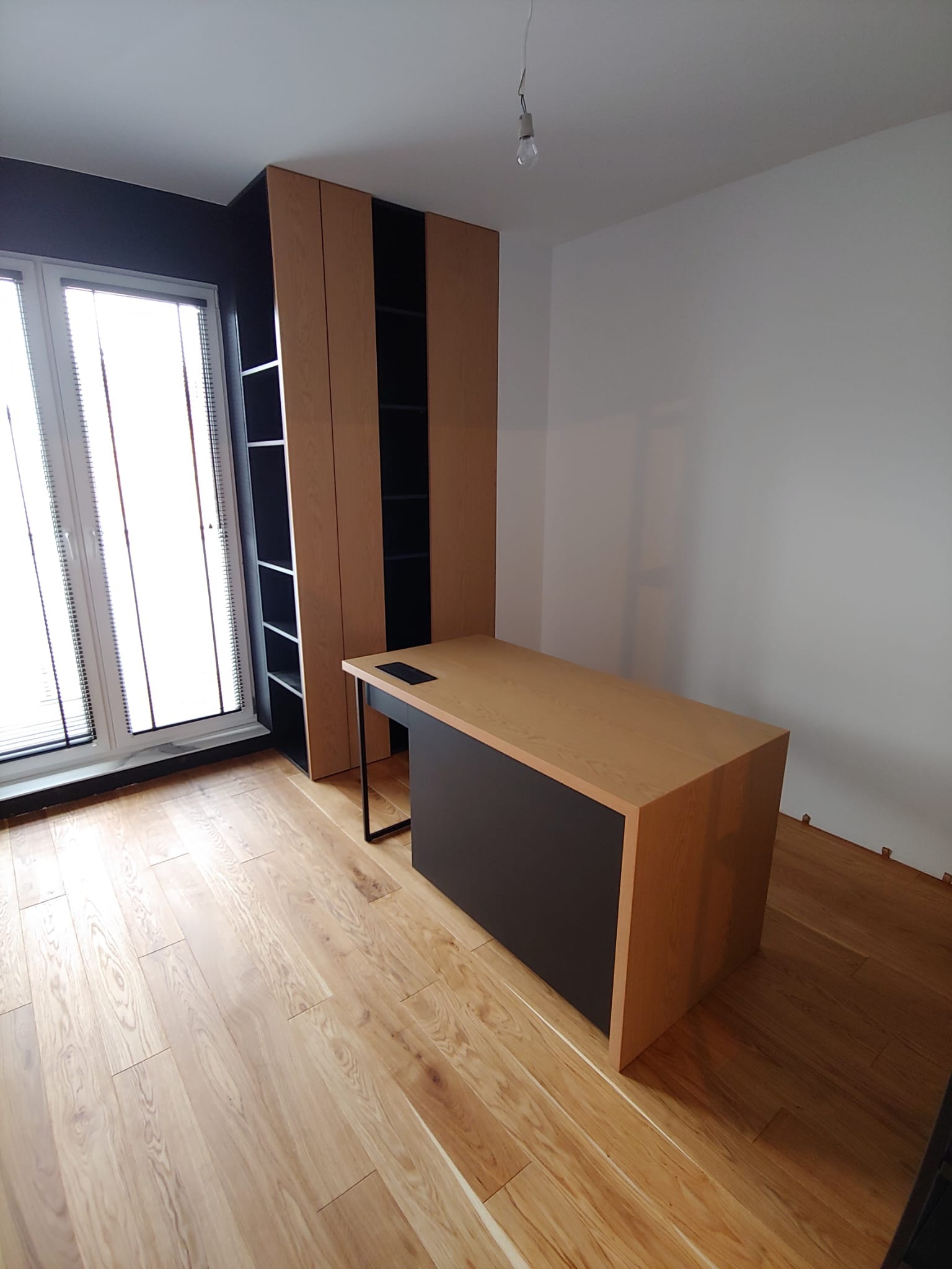 Pokój z biurkiem z blatem drewnianym i czarnymi nogami, obok regał z półkami, podłoga drewniana, okno z żaluzjami.