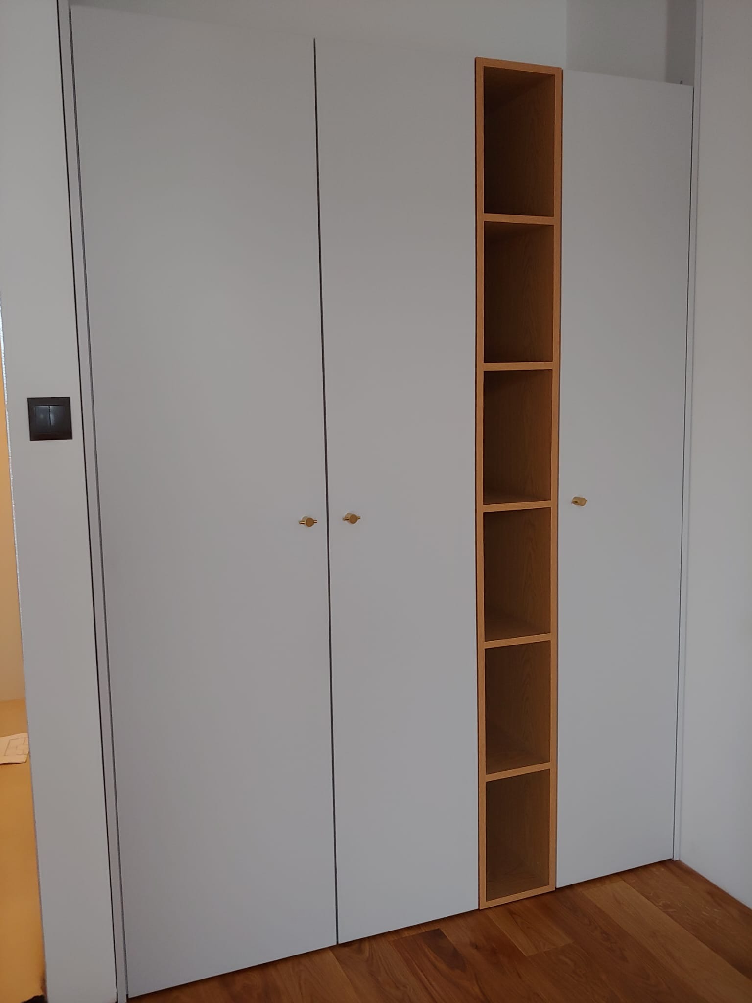 Biała szafa wnękowa z jasnymi, okrągłymi uchwytami i otwartą, drewnianą wnęką z półkami, widoczna na tle drewnianej podłogi.