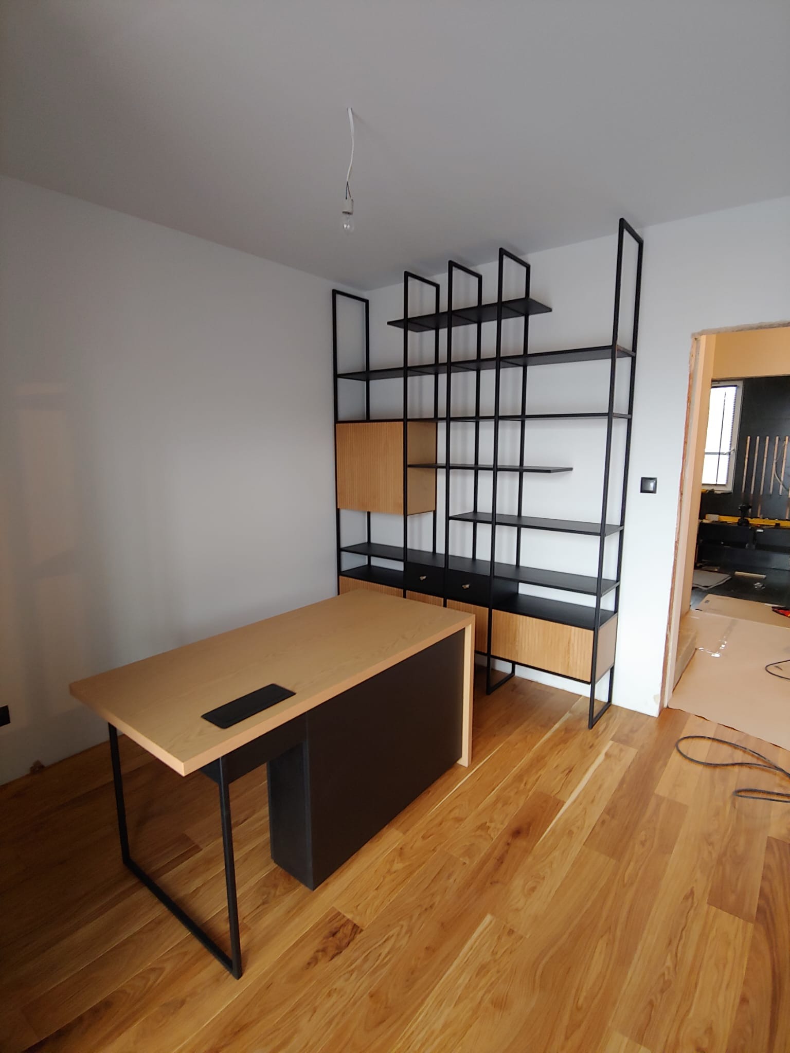 Nowoczesny pokój z biurkiem z blatem w odcieniach drewna i czarną podstawą oraz regałem z czarnymi metalowymi ramami i drewnianymi elementami, parkiet na podłodze, widok na kolejne pomieszczenie...