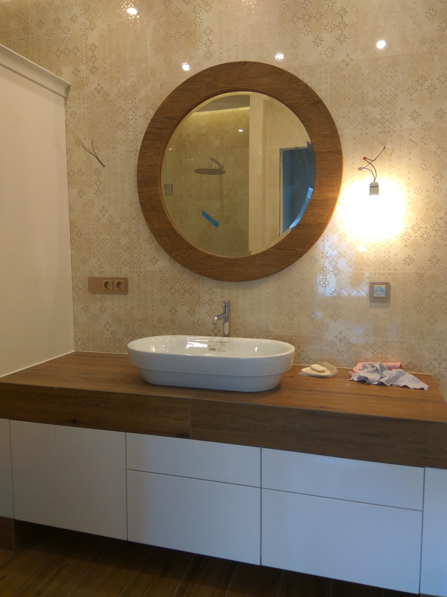 Łazienka z okrągłym lustrem w drewnianej ramie, białą umywalką nablatową i blatem z drewna na białych szafkach; widoczne gniazdka elektryczne w ścianie z beżowych płytek z kwiatowym wzorem.