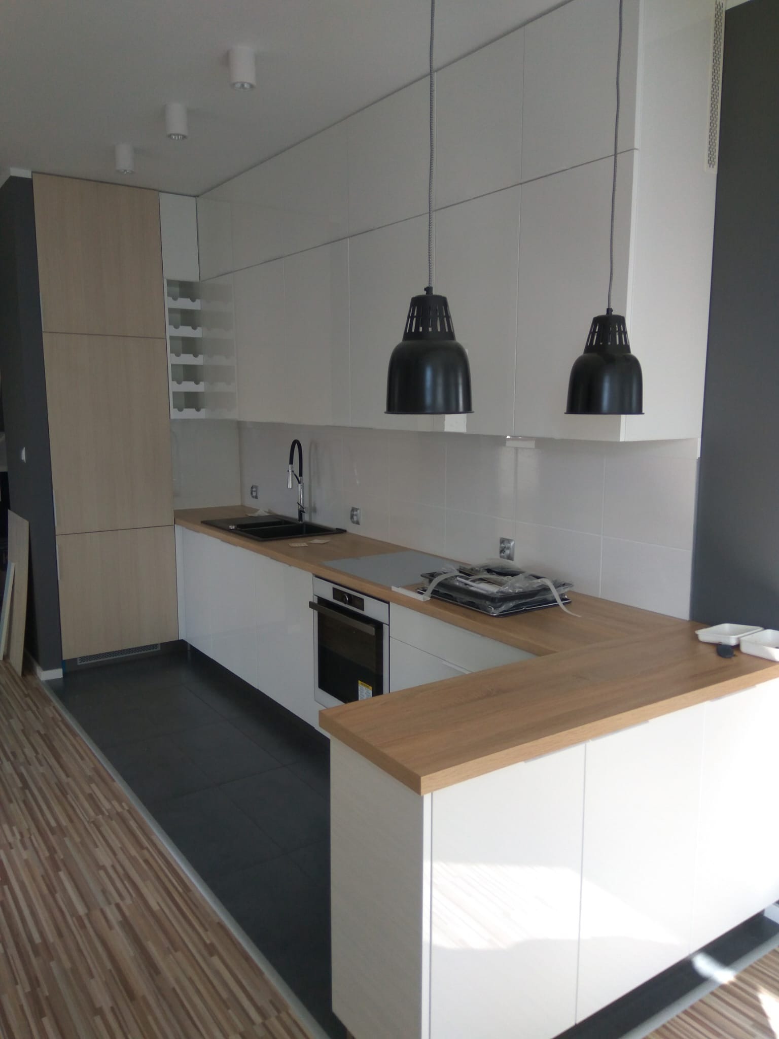 Nowoczesna kuchnia z białymi frontami i drewnianym blatem, czarnym zlewem i wiszącymi lampami, widok na fragment podłogi z płytek i paneli.