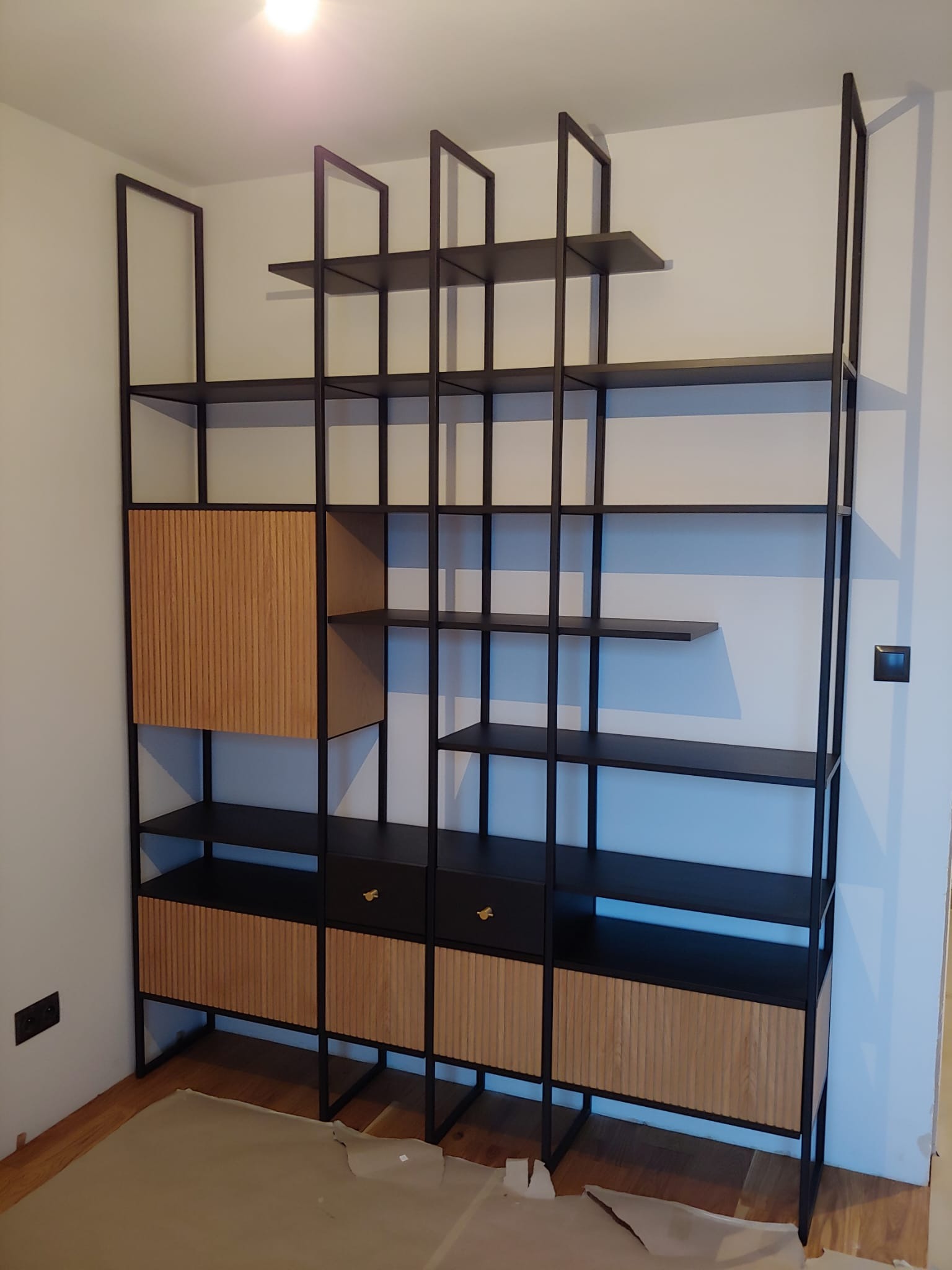 Regal z czarną metalową konstrukcją i drewnianymi elementami w odcieniach brązu, zlokalizowany we Wrocławiu, na podłodze widoczne zabezpieczenia.