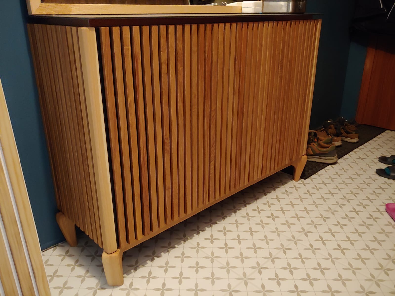 Drewniana szafka z ryflowanymi drzwiami, stojąca na wzorzystej podłodze, z widocznymi butami w tle.