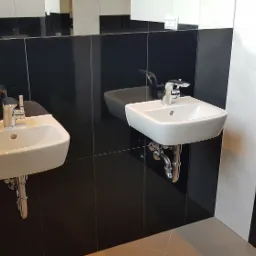 Dwie białe umywalki zamontowane na czarno-białej ścianie z płytek, chromowane baterie i syfony, widoczne odbicie osoby robiącej zdjęcie.