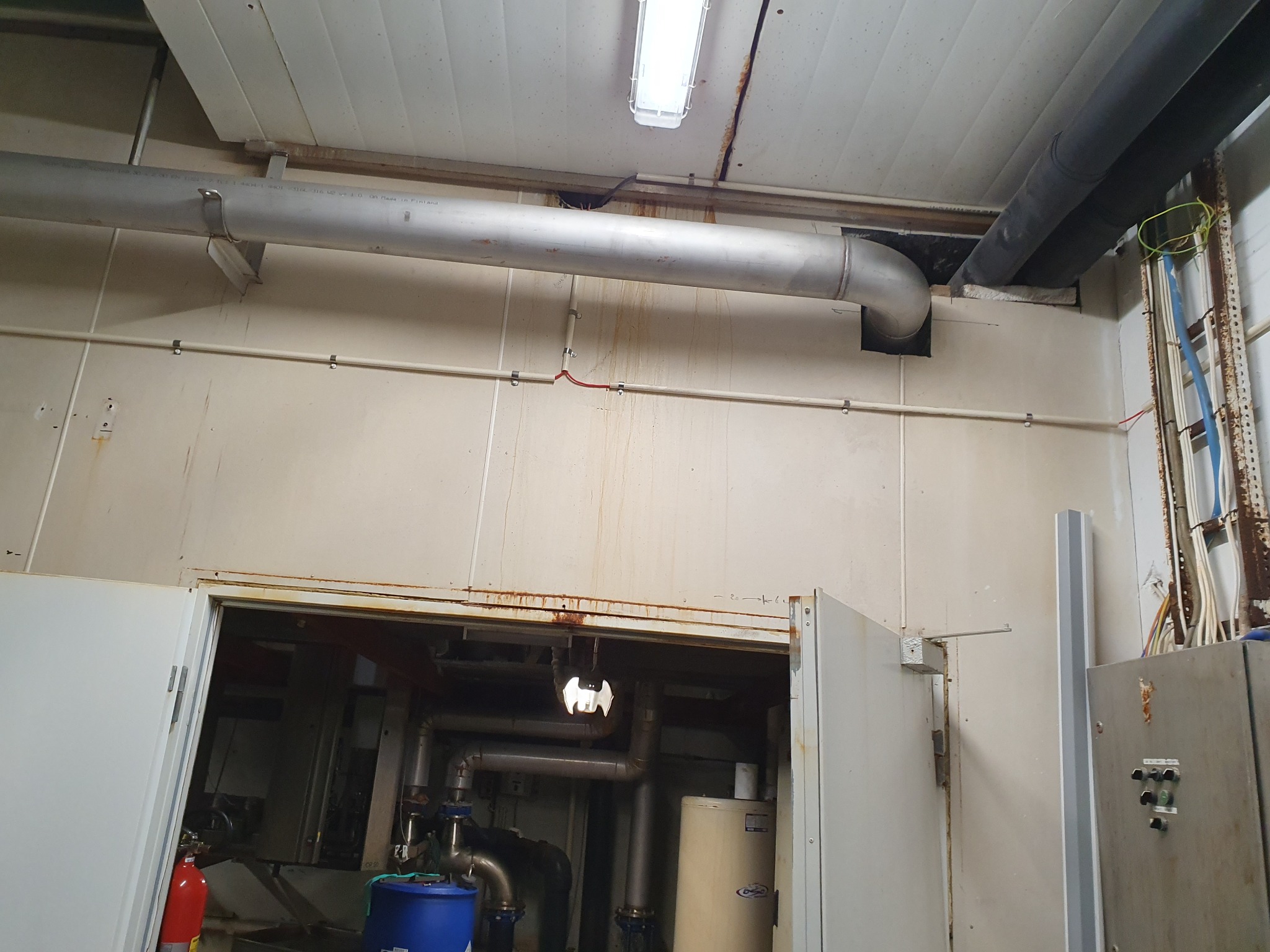 Wnętrze hali przemysłowej z widocznymi rurami instalacyjnymi pod sufitem, oświetleniem i częściowo otwartymi drzwiami do pomieszczenia z zbiornikami i rurami.