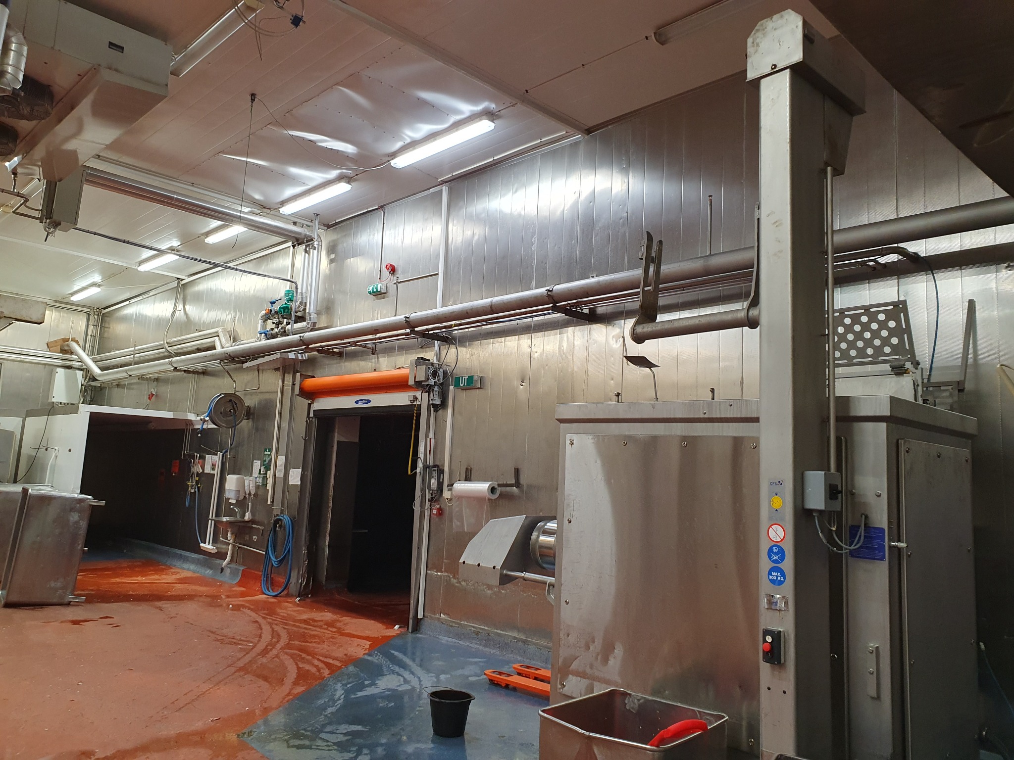 Wnętrze przemysłowej chłodni z metalowymi ścianami i podłogą w odcieniach czerwieni i szarości, widoczne rury, armatura, urządzenia chłodnicze oraz otwarte drzwi do komory chłodniczej.