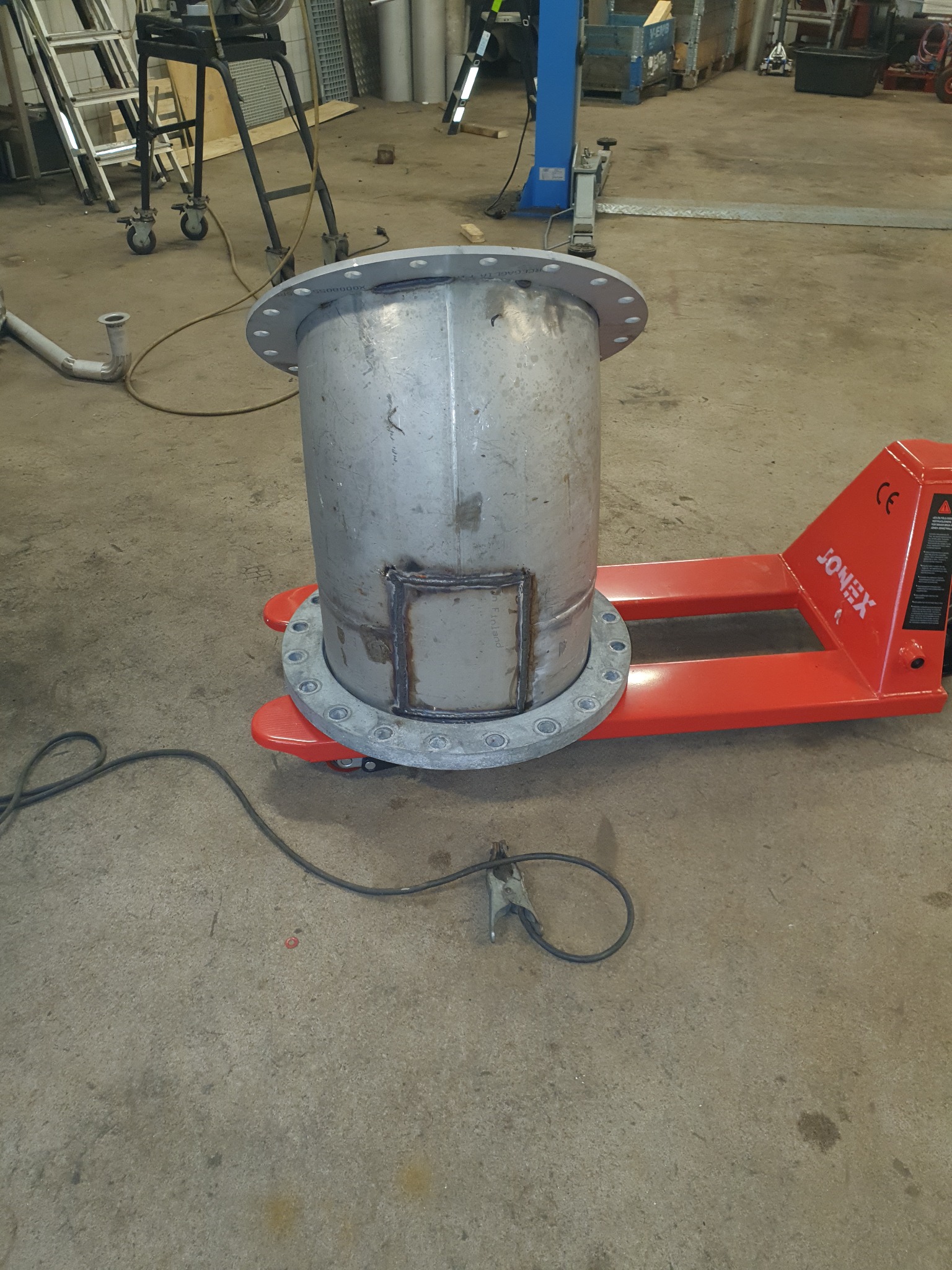 Metalowy cylinder z przyspawaną klapą i kołnierzami na wózku paletowym, widoczny przewód spawalniczy na betonowej podłodze warsztatu.