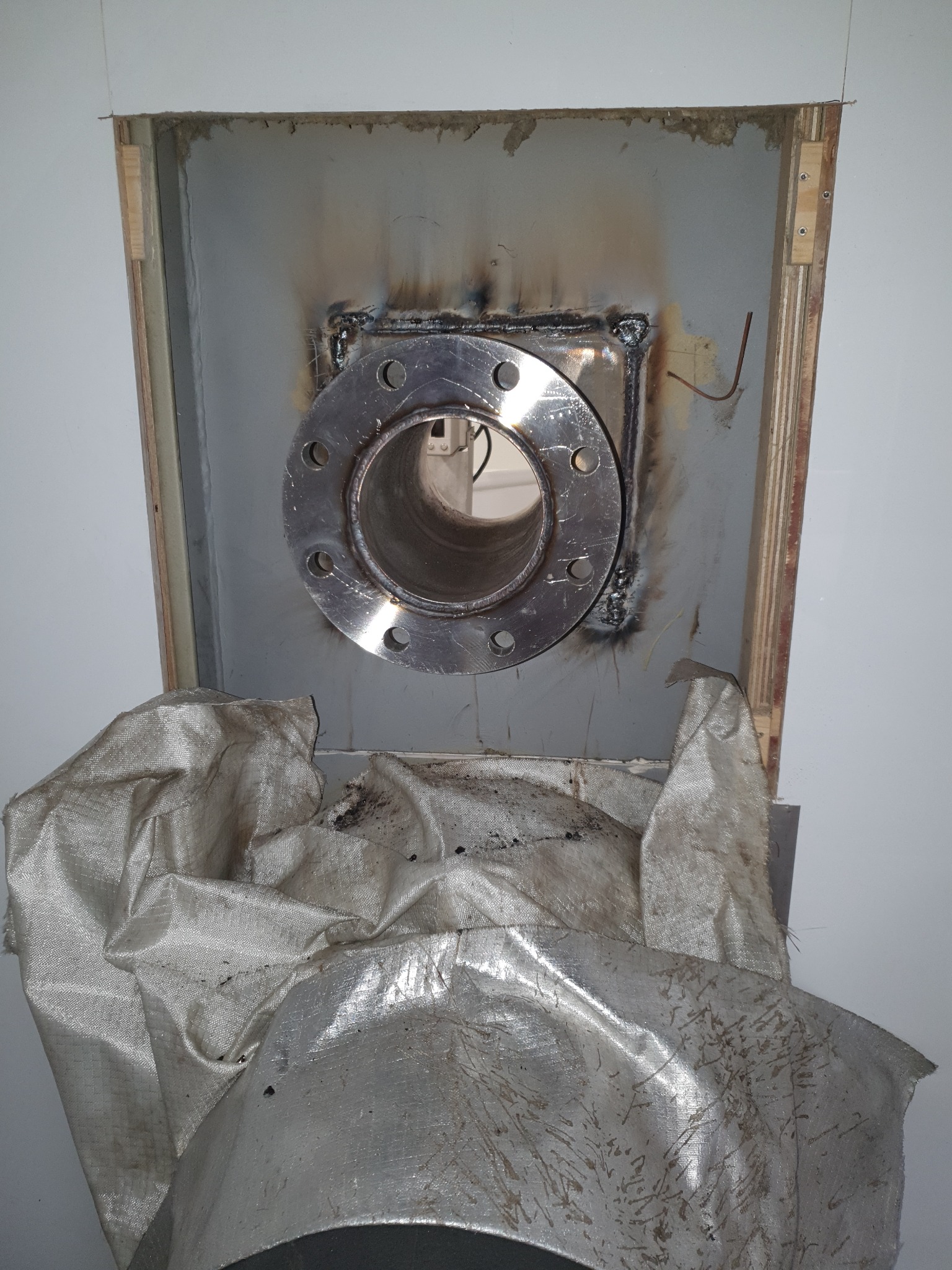 Metalowy kołnierz zespawany z blachą w otworze ściany, widoczne ślady spawania i zabezpieczająca folia.