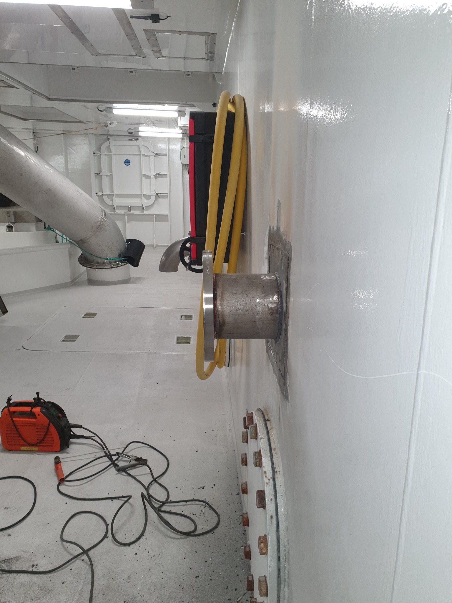 Wnętrze przemysłowe z widocznym spawarką, metalowym rurociągiem i żółtym wężem, ukazujące prace związane z instalacją lub naprawą.