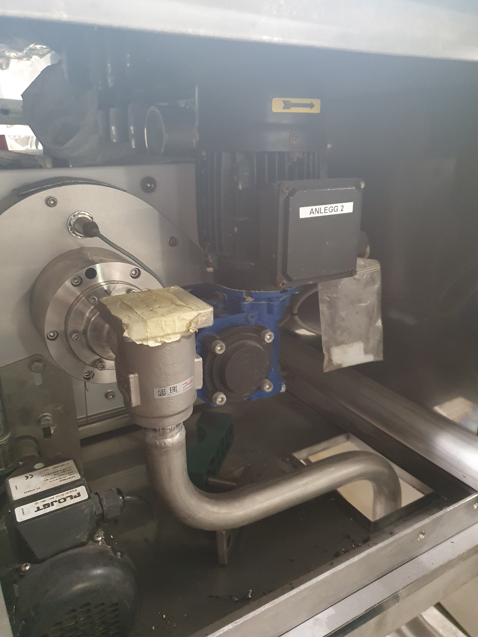 Wnętrze maszyny z metalowymi rurami, silnikiem i osprzętem, widoczna tabliczka 'ANLEGG 2' i żółta strzałka.