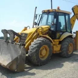 Żółta koparko-ładowarka JCB SiteMaster na placu budowy, widoczna z przodu, z podniesioną łyżką i wysuniętym ramieniem kopiącym w tle, kolejna maszyna w oddali.