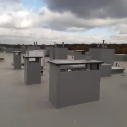 Szary, płaski dach z licznymi kominami wentylacyjnymi o kwadratowym przekroju, widok z góry na tle pochmurnego nieba i drzew w oddali.