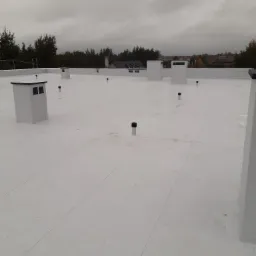 Biały dach płaski z obróbkami blacharskimi i kominami wentylacyjnymi na tle pochmurnego nieba i zabudowań.