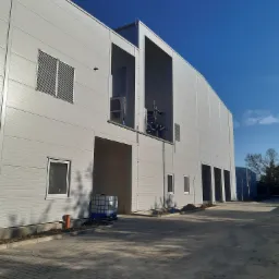 Nowo wybudowany magazyn z szarej blachy trapezowej, widoczne okna, kratki wentylacyjne oraz instalacje na zewnątrz budynku, niebieskie niebo w tle.