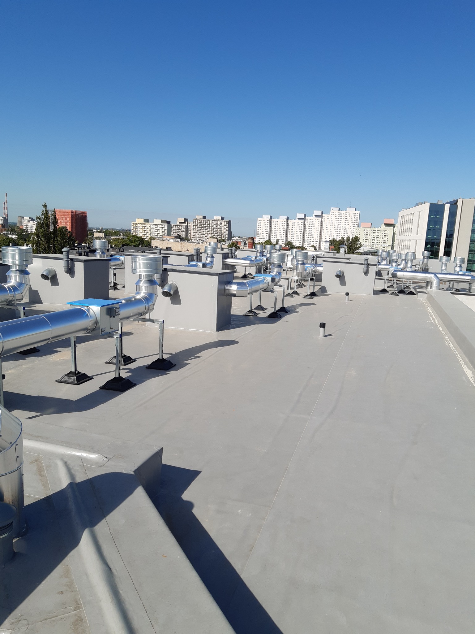 Szary płaski dach z licznymi systemami wentylacyjnymi i klimatyzacyjnymi, w tle zabudowa miejska pod błękitnym niebem.