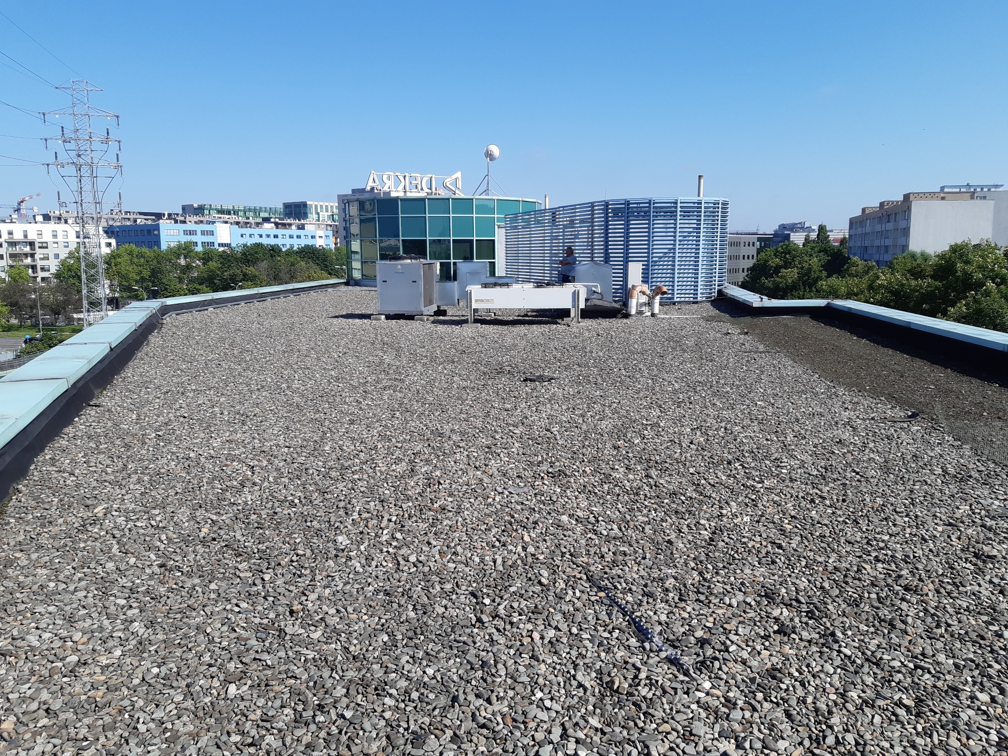 Płaski dach pokryty drobnym żwirem z widocznymi elementami wentylacji i klimatyzacji na tle zabudowy miejskiej w słoneczny dzień.