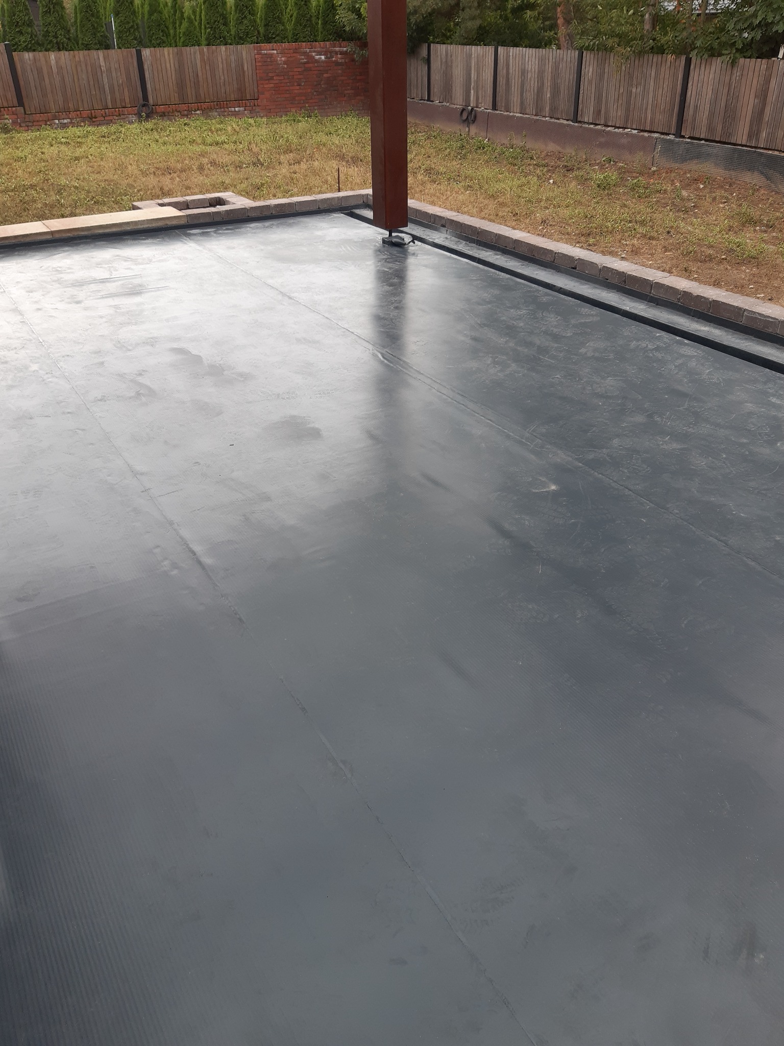 Nowa, szara, płaska powierzchnia dachu pokryta papą termozgrzewalną, z obrzeżem z cegieł i elementami konstrukcyjnymi w tle.