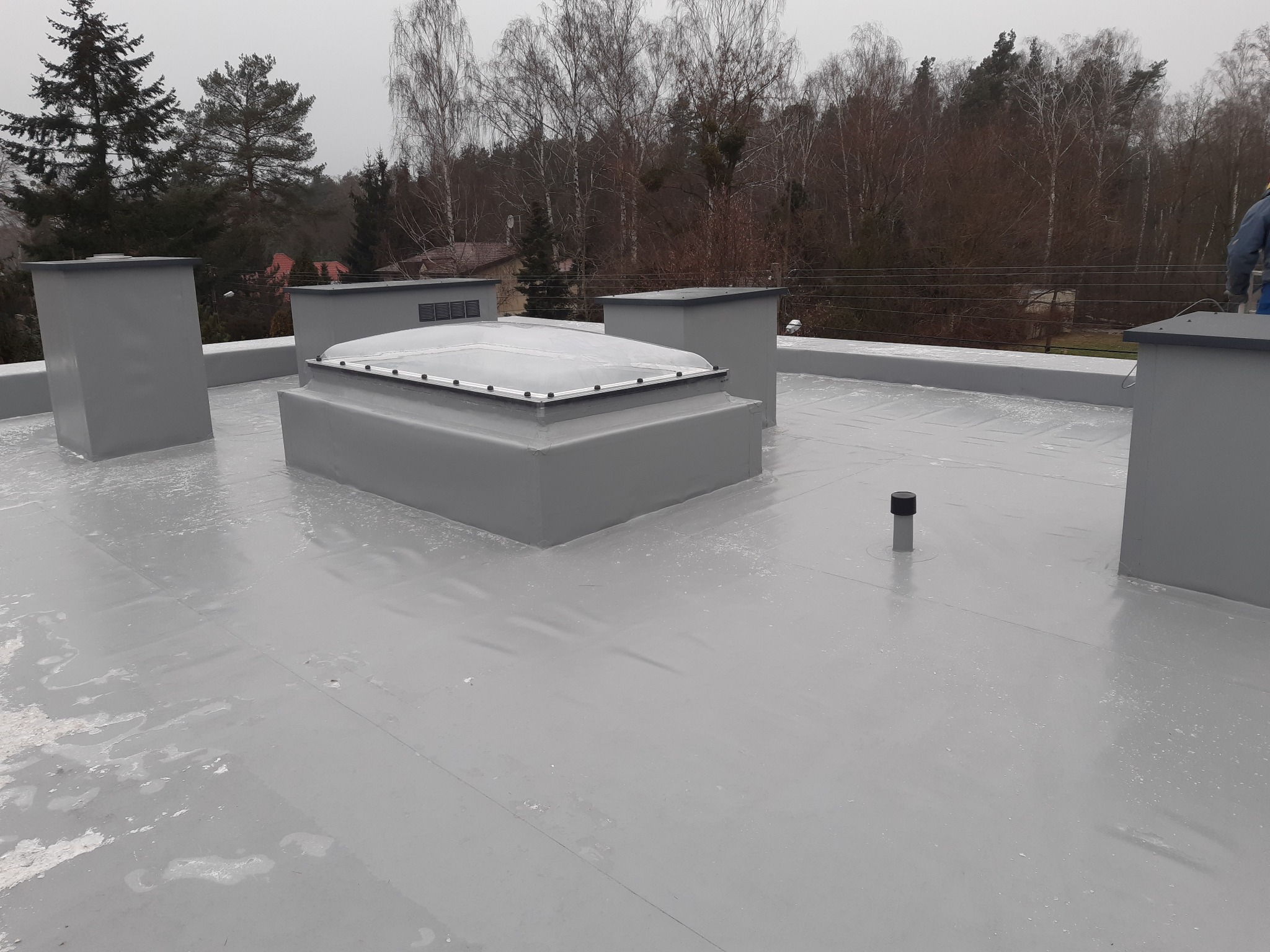 Szary dach pokryty papą termozgrzewalną z czterema kominami wentylacyjnymi i świetlikiem dachowym na tle drzew