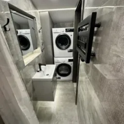 Wąska łazienka w szarościach: pralka i suszarka w kolumnie, umywalka z szafką, lustro z wieszakami oraz grzejnik drabinkowy. Nowoczesne oświetlenie sufitowe.