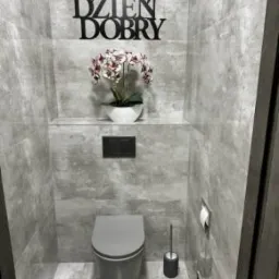 Szara, nowoczesna toaleta z płytkami imitującymi beton, dekoracją 'Dzień Dobry' i kwiatami. Minimalistyczny design, oświetlenie sufitowe.